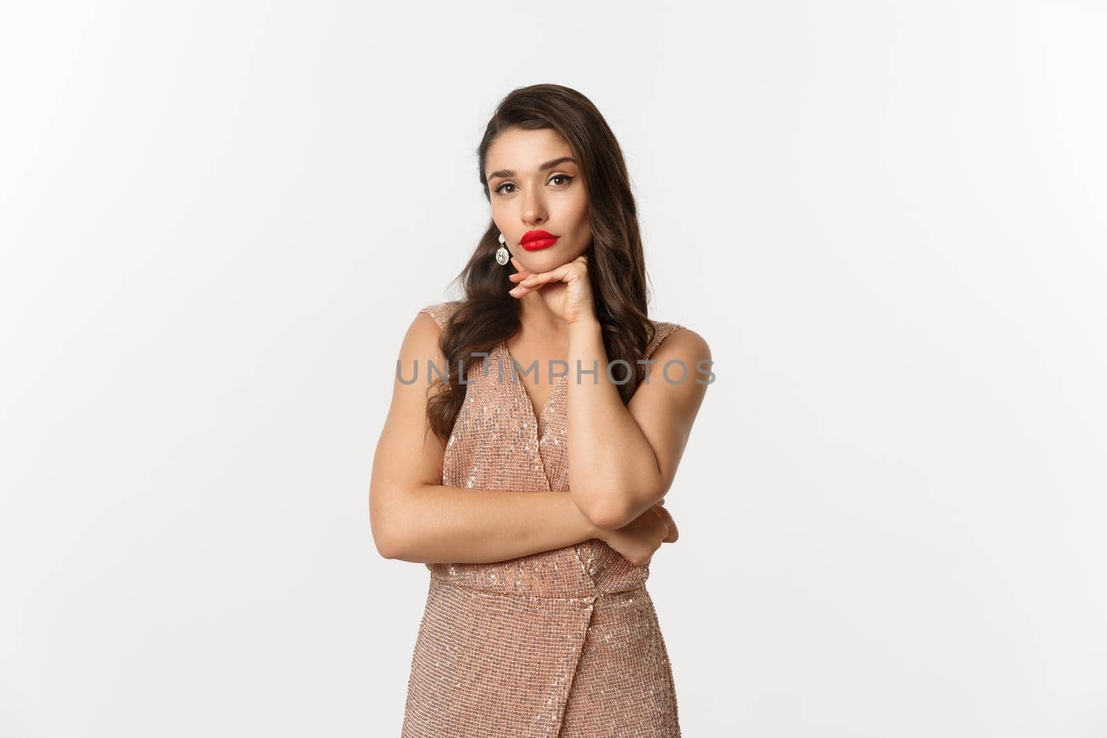 Celebration and party concept. Beautiful young female model in glamour dress looking thoughtful at camera, staring and making decision, standing over white background.