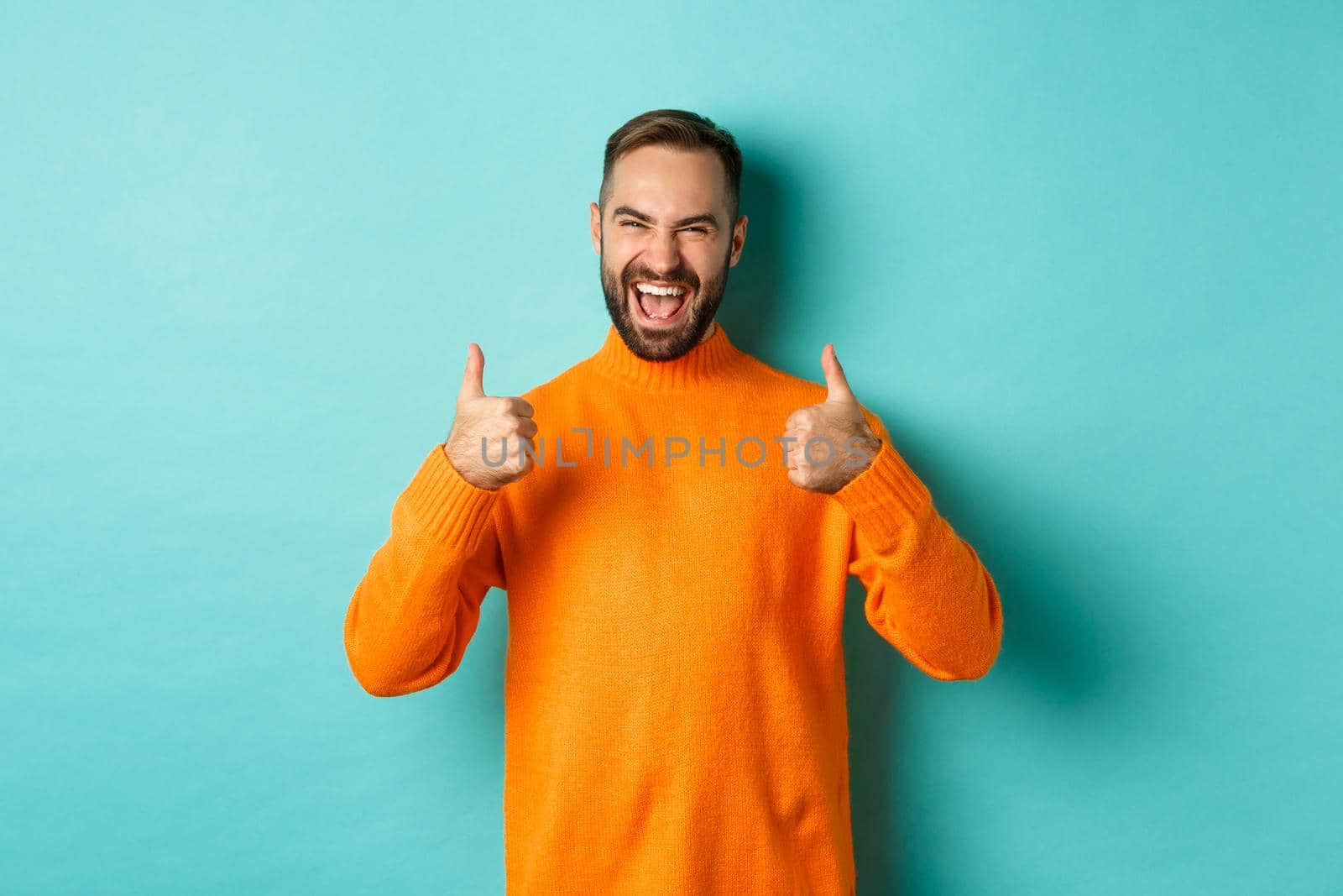 Super good. Satisfied happy man showing thumbs up, agree with you, praise excellent work, looking pleased, standing in orange sweater against light blue background.