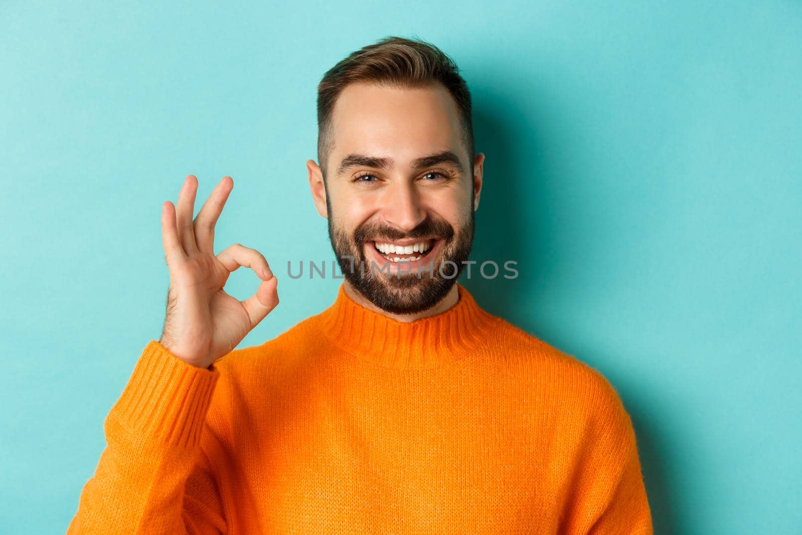 Close-up of assertive young man assuring everything ok, showing okay sign and smiling, yes or positive answer, standing over light blue background.