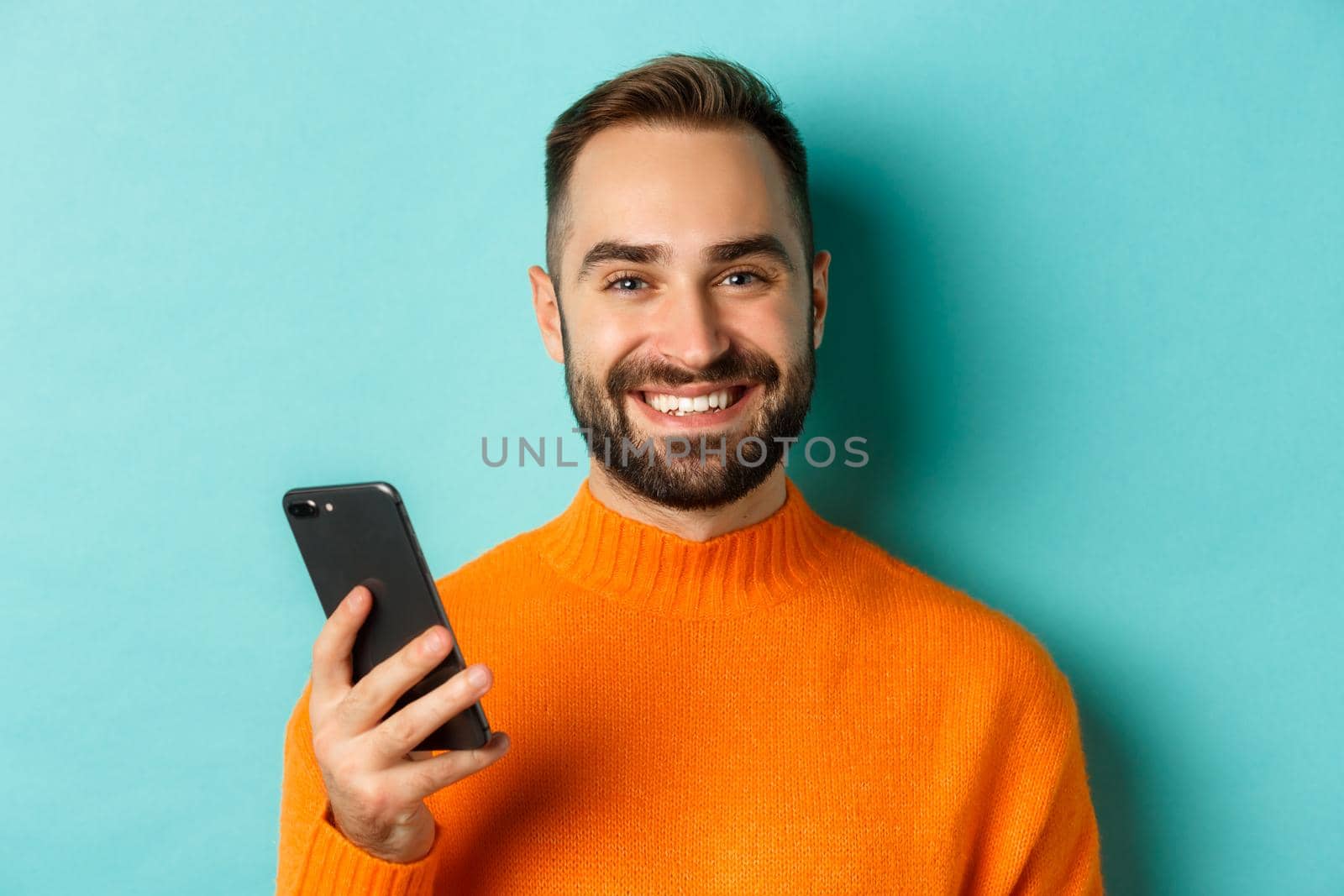 Close-up of happy handsome man writing message on mobile phone, holding smartphone and smiling, standing against turquoise background.
