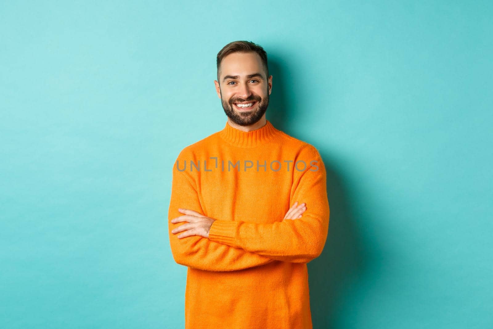 Handsome bearded male model in orange sweater, looking confident and smiling, cross arms on chest, standing over turquoise background.