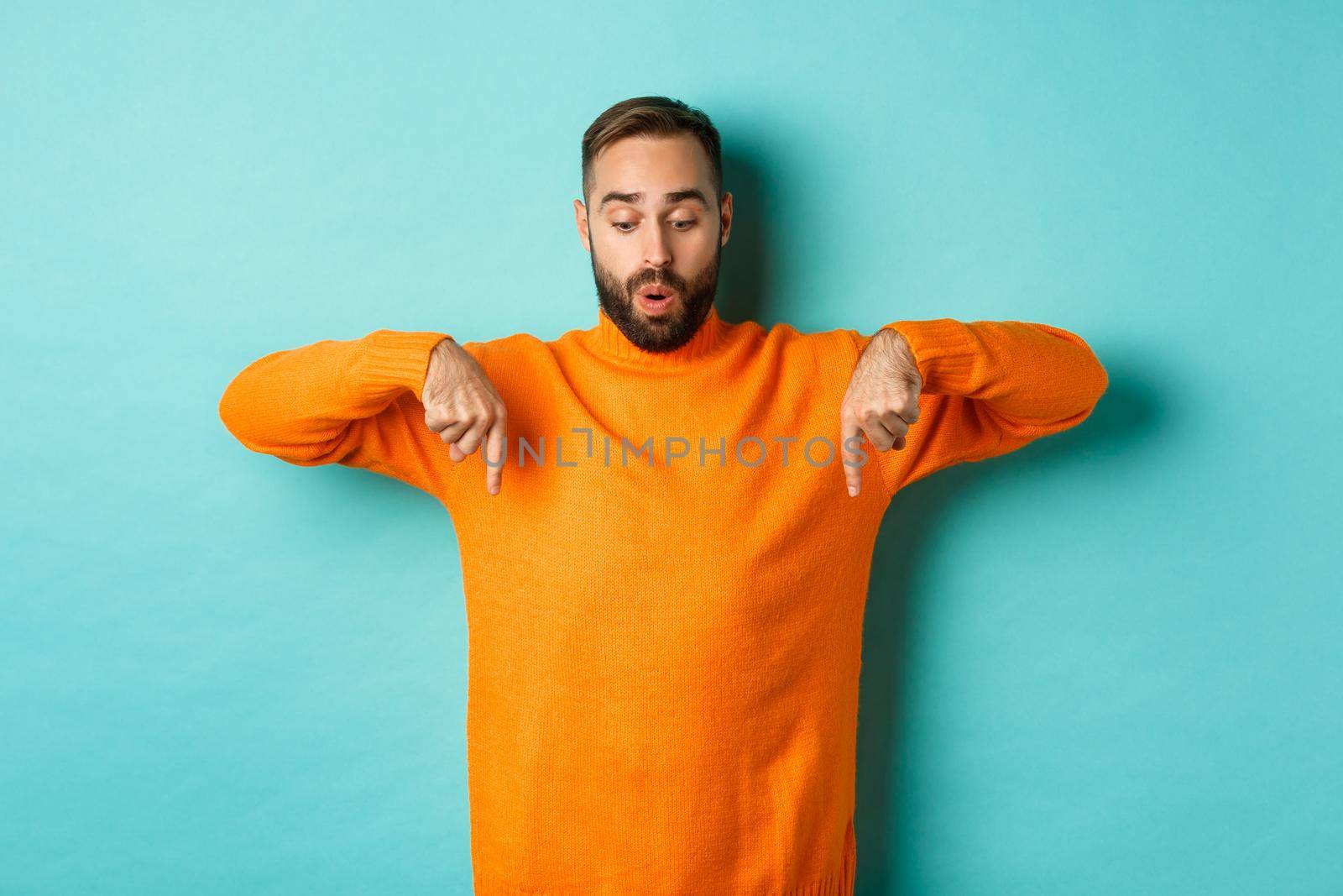 Amazed guy in orange sweater, pointing fingers down and looking with interest at promo, holiday discounts, standing over turquoise background.