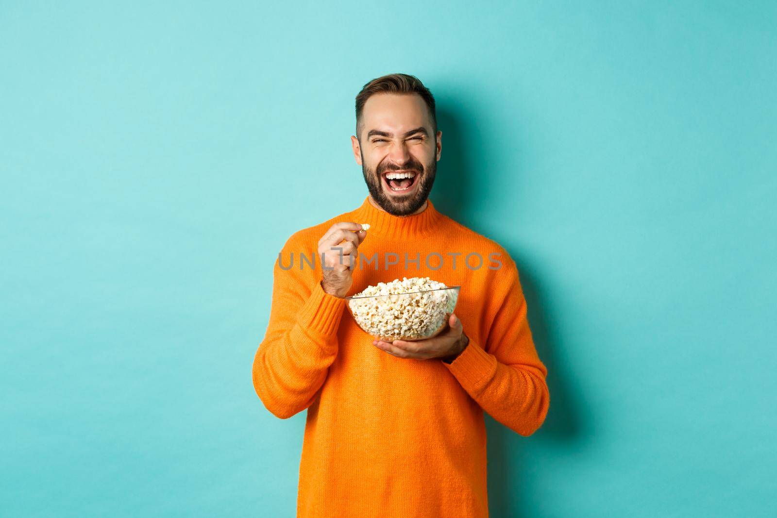 Excited young man watching interesting movie on tv screen, eating popcorn and looking amazed, blue background.