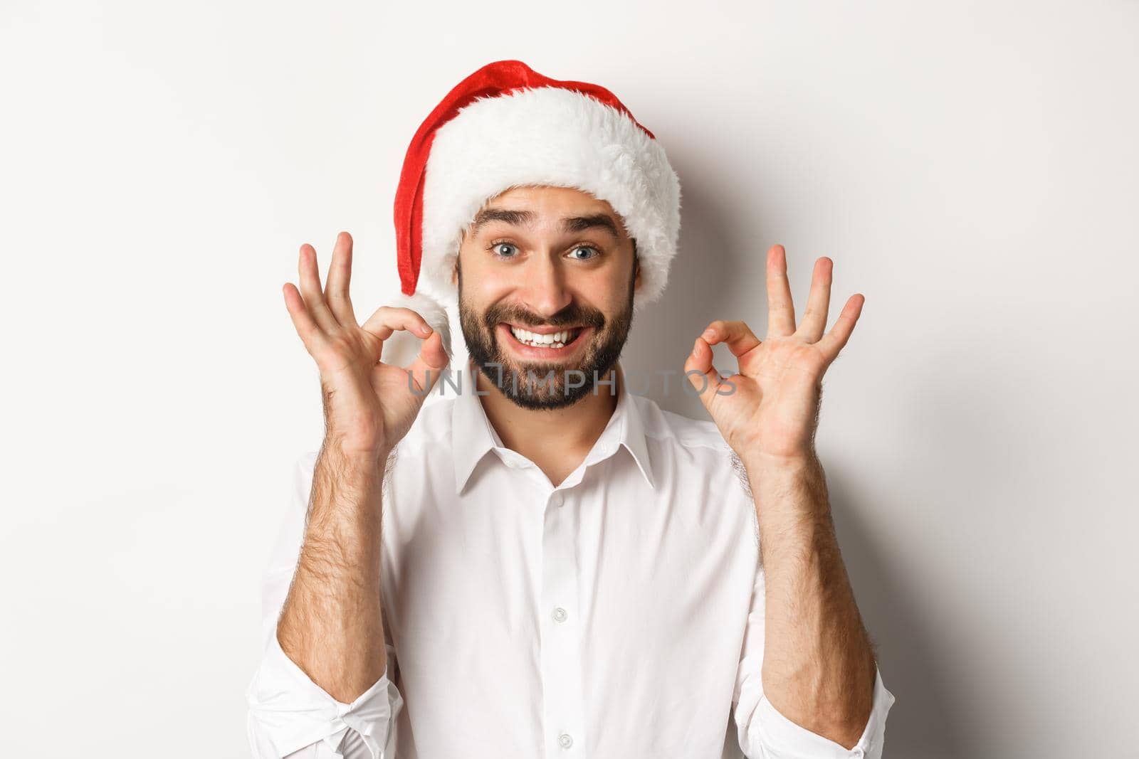 Party, winter holidays and celebration concept. Joyful man enjoying christmas and showing okay sign, smiling satisfied, wearing santa hat, white background.
