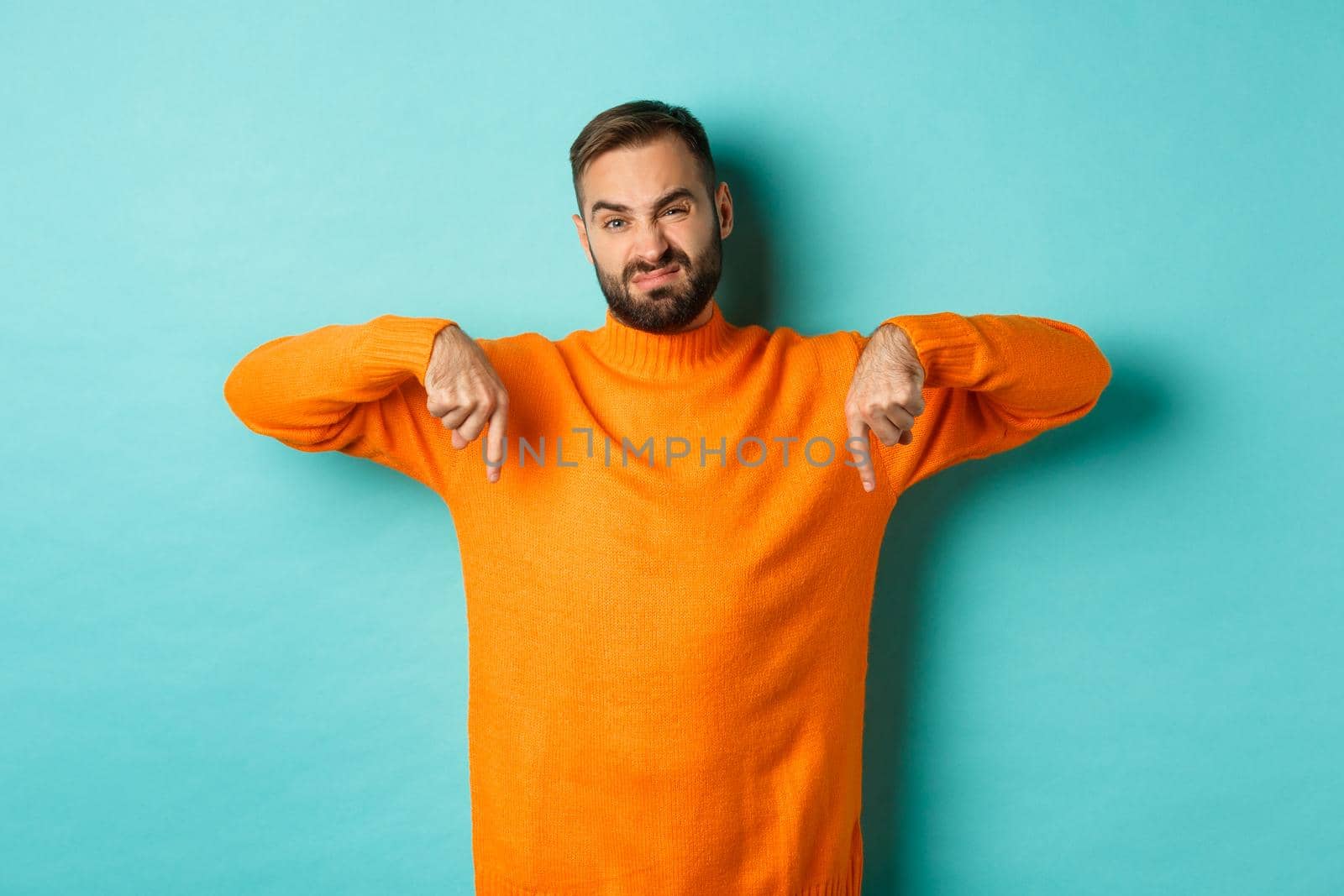 Displeased and skeptical handsome man pointing fingers down, showing bad product, dislike store or advertisement, grimacing disappointed, standing over turquoise background.