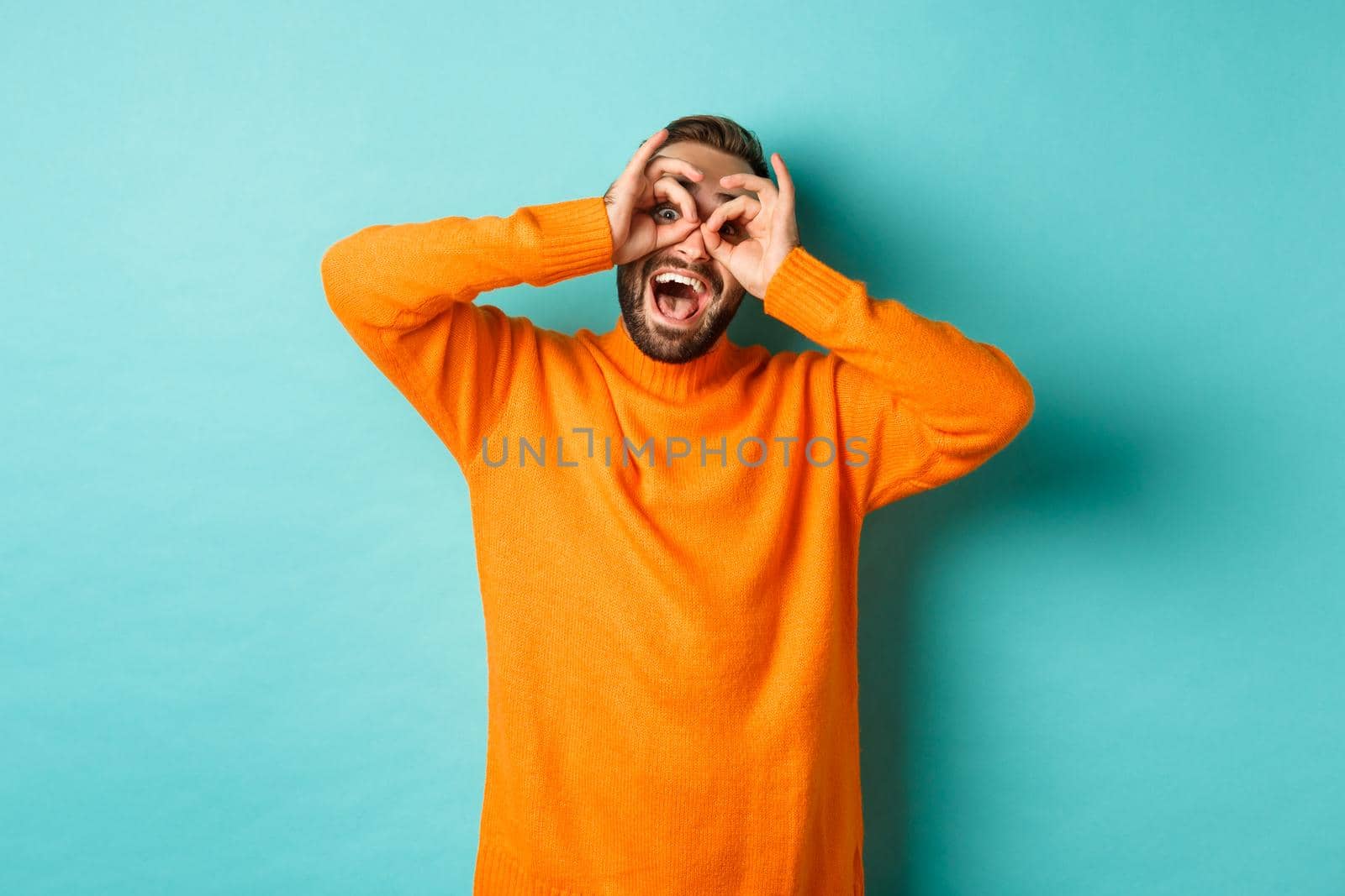 Image of funny and happy guy holding hands on eyes, looking through fake glasses, smiling amused, standing in orange sweater against turquoise background.