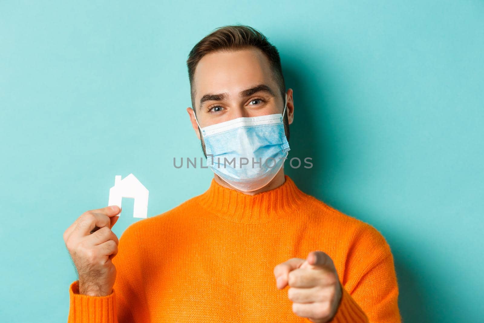 Real estate and coronavirus pandemic concept. Close-up of adult man in medical mask pointing at you and showing small paper house, standing over light blue background.