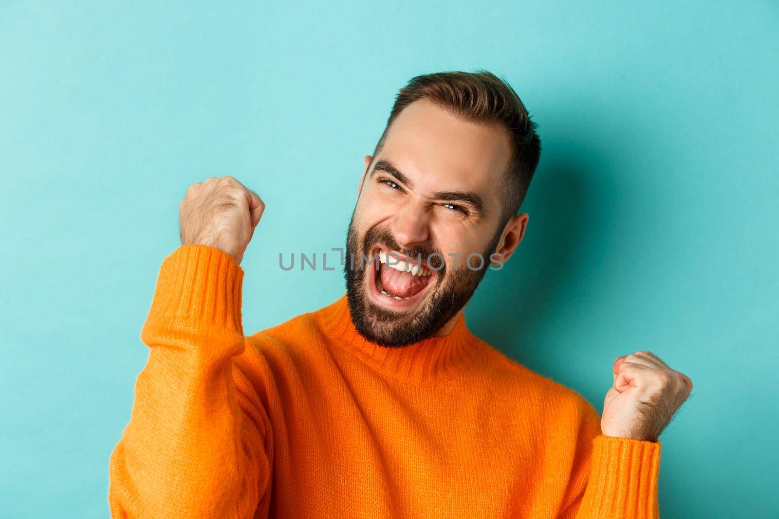 Image of handsome relieved man feeling satisfaction, rejoicing of winning or achievement, making fist pump and saying yes, standing over turquoise background.