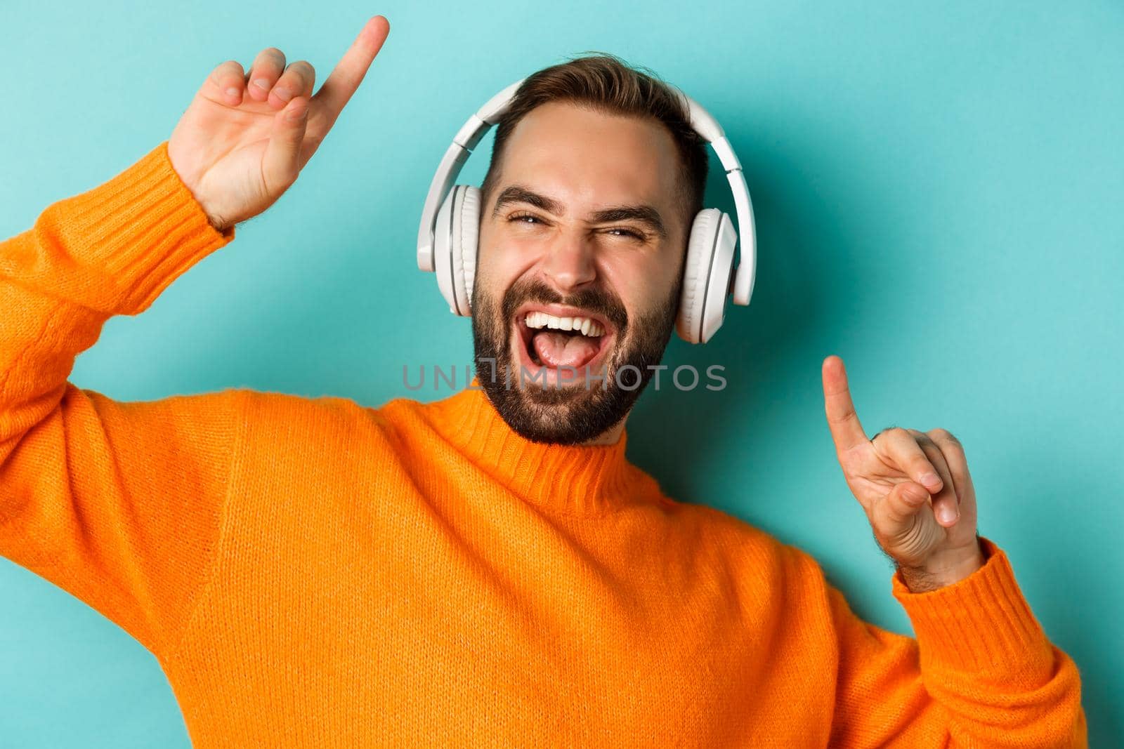Close-up of handsome modern man listening music in headphones, standing in orange sweater over turquoise background.