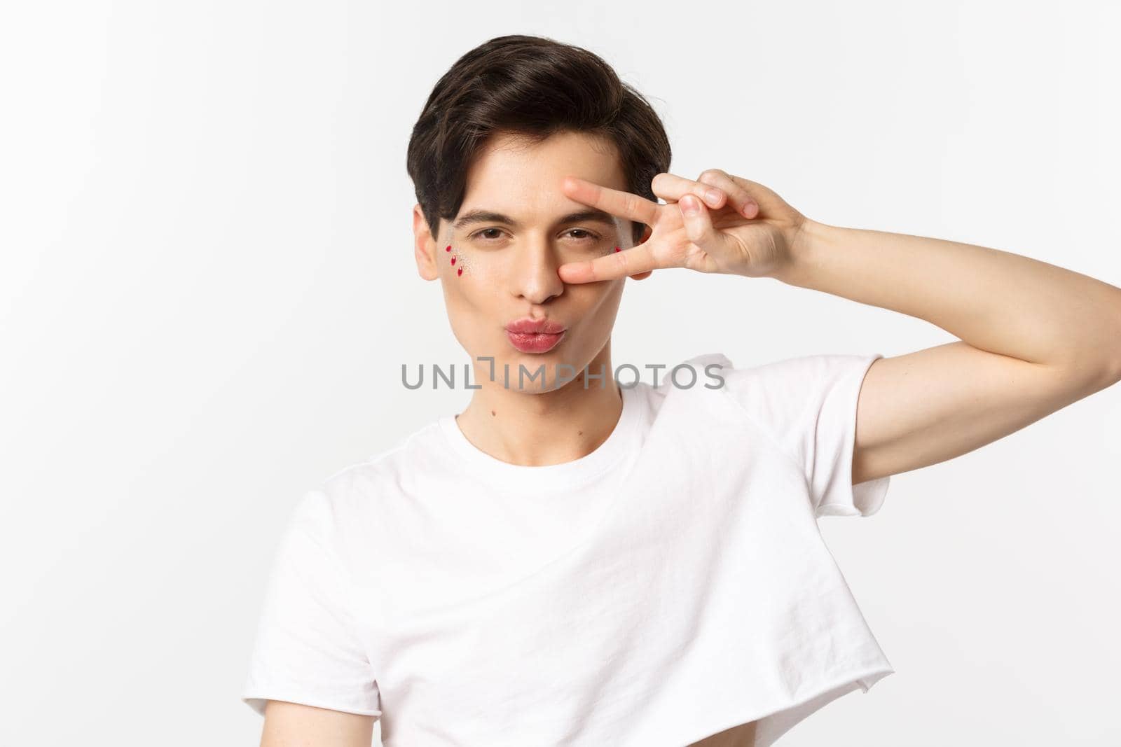 People, lgbtq community and lifestyle concept. Close-up of sassy and beautiful gay man with glitter on face, pucker lips in kiss and showing peace sign, white background.
