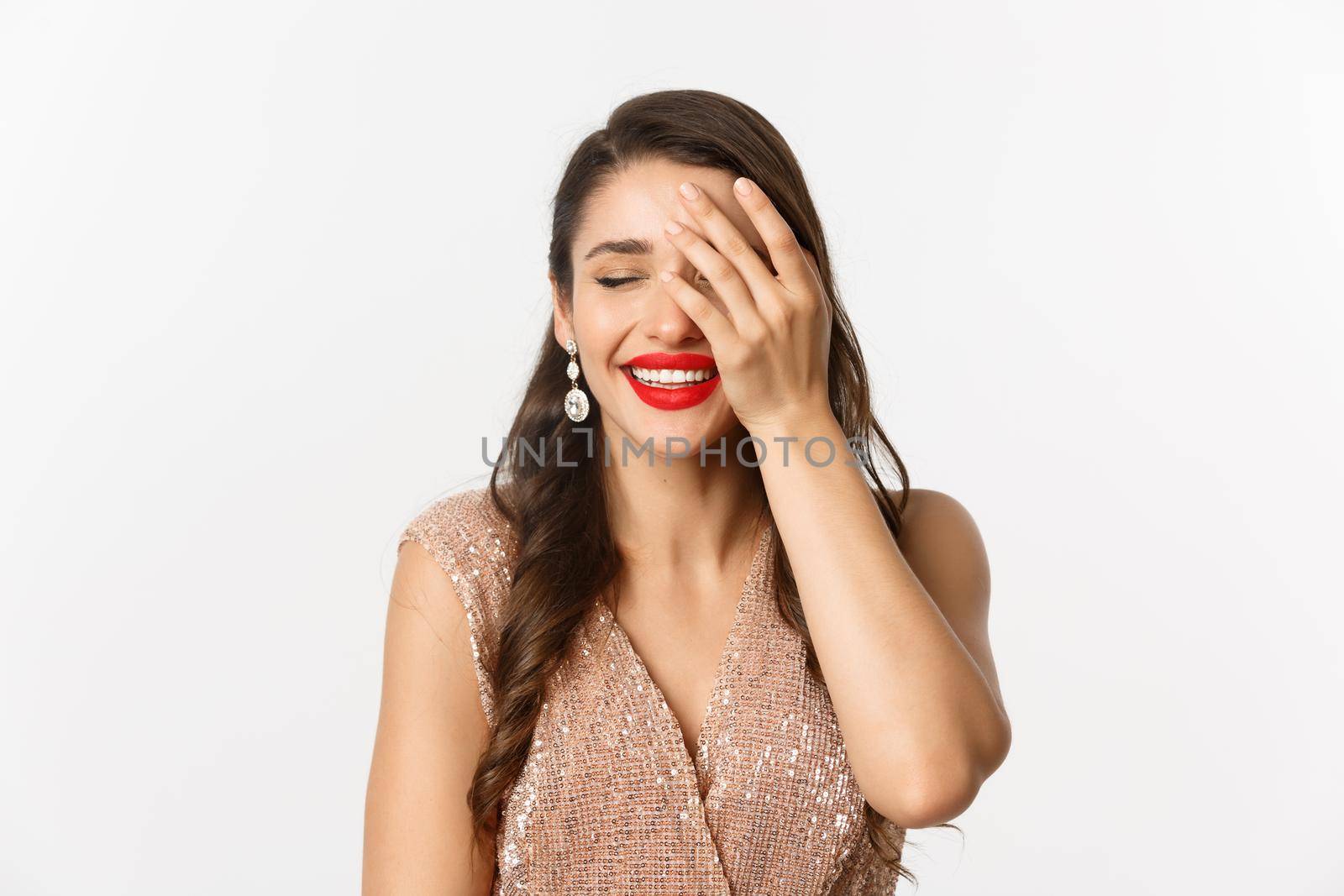 Concept of New Year celebration and winter holidays. Fabulous woman with red lips, wearing party dress, laughing and looking delighted, standing over white background.