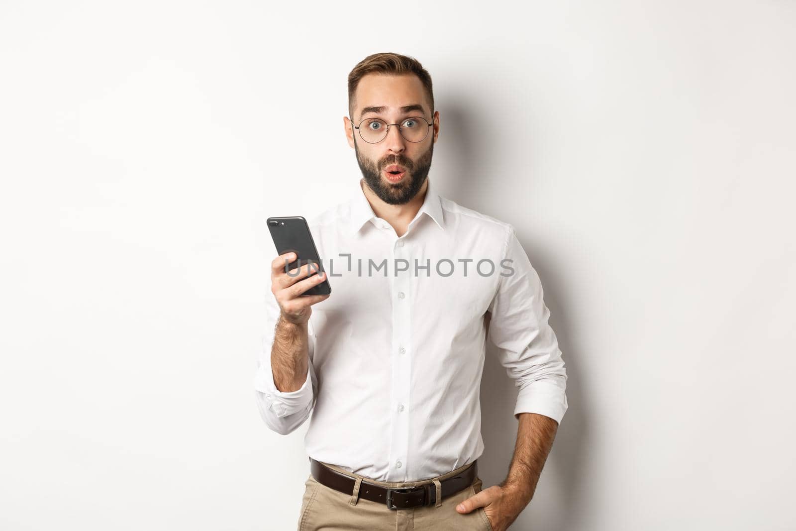 Surprised handsome man reading interesting info online, holding smartphone, standing over white background.