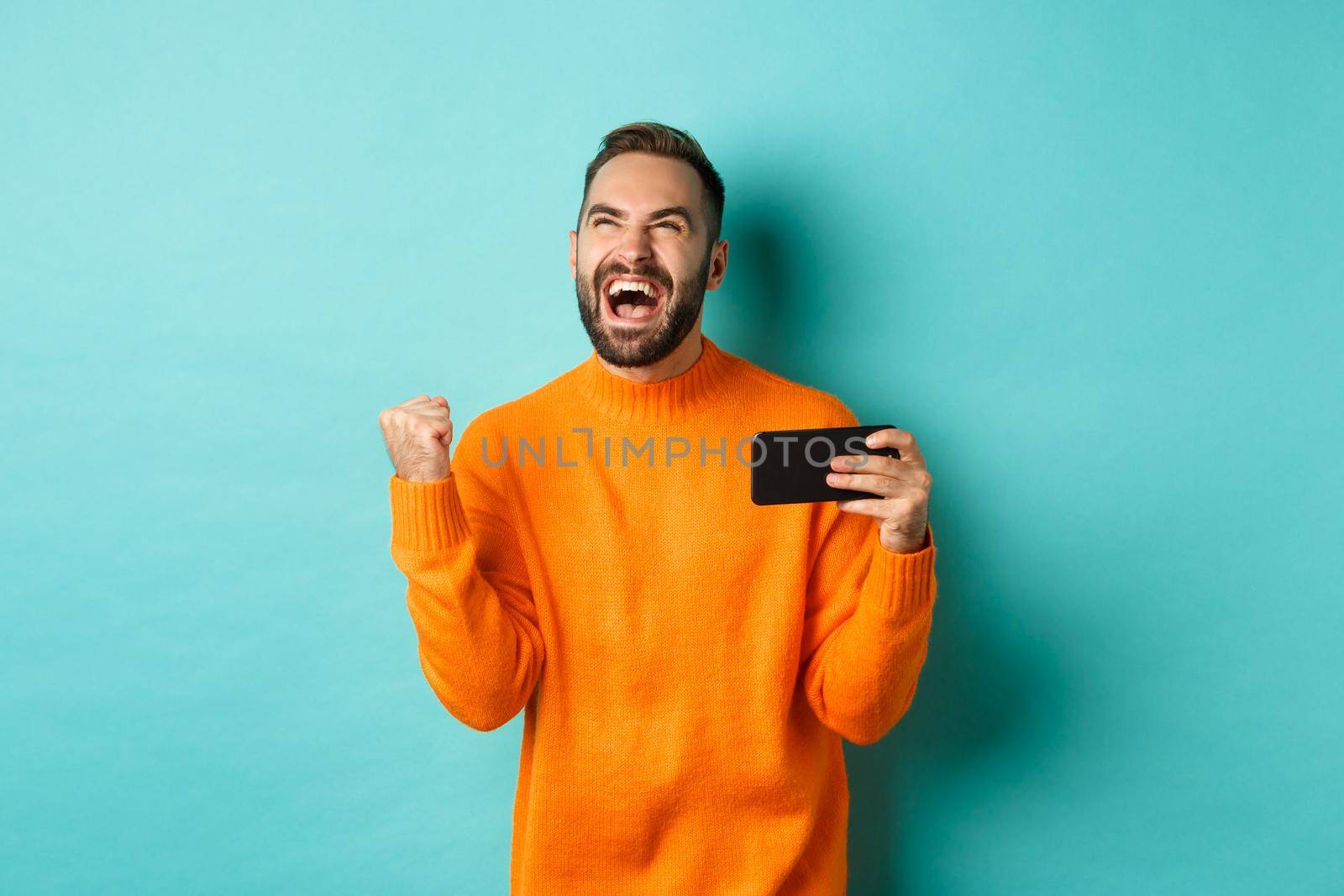 Image of man winning on mobile phone, making fist pump and rejoicing of victory, holding smartphone with satisfied face, turquoise background.