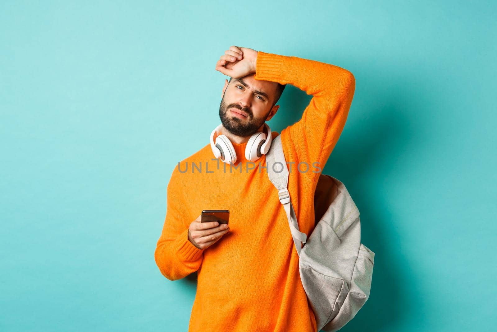 Tired guy with headphones and backpack, wiping sweat off forehead with exhausted face, using mobile phone, standing over turquoise background.