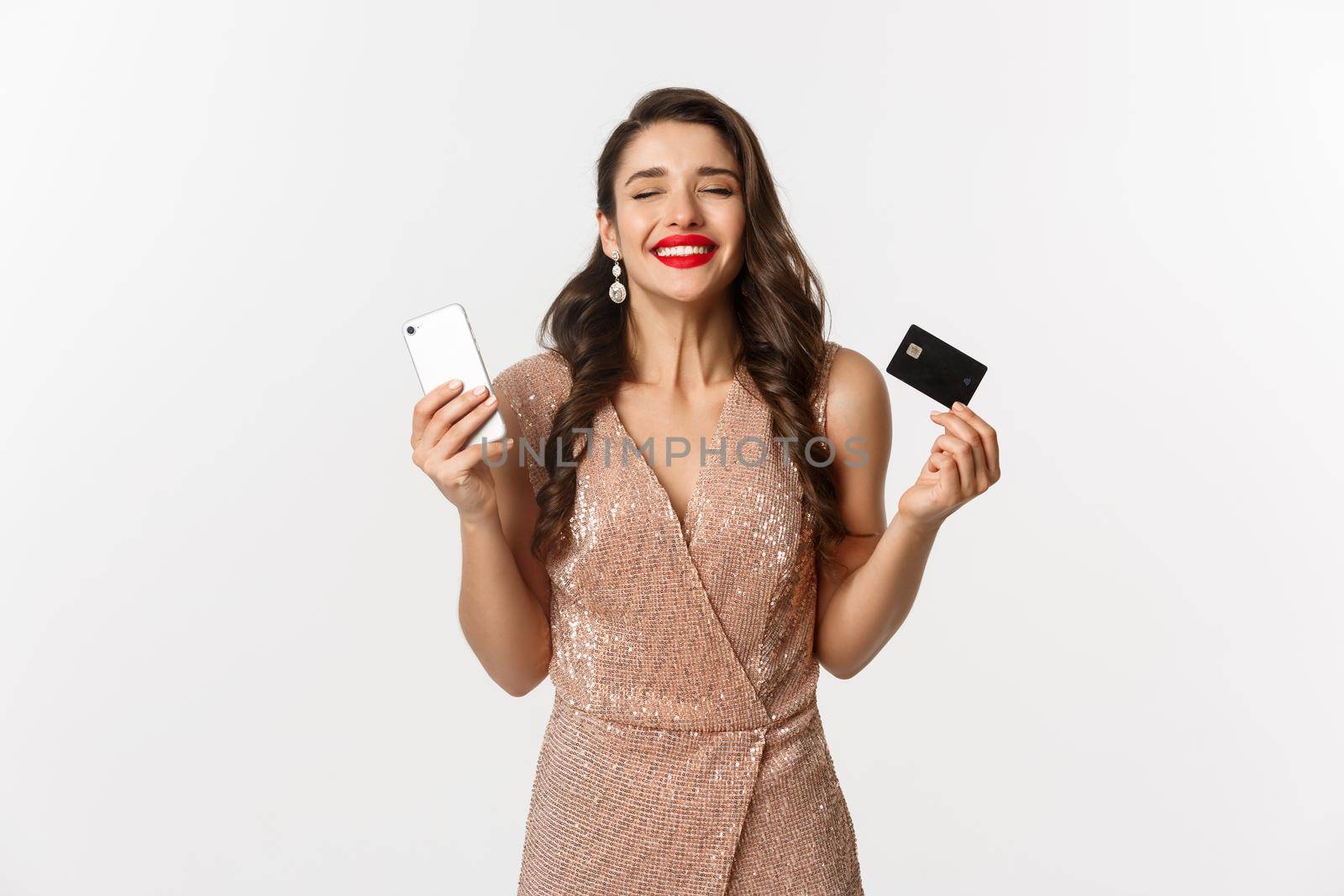 Online shopping and holidays concept. Satisfied and happy woman in elegant dress smiling, using credit card and mobile phone, standing over white background.