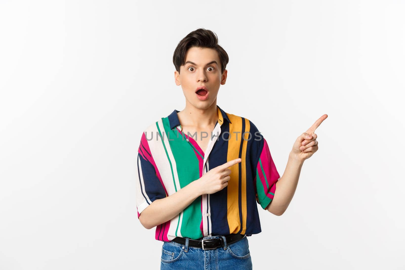 Impressed young man showing advertisement, open mouth with disbelief and pointing at upper right corner, standing against white background.