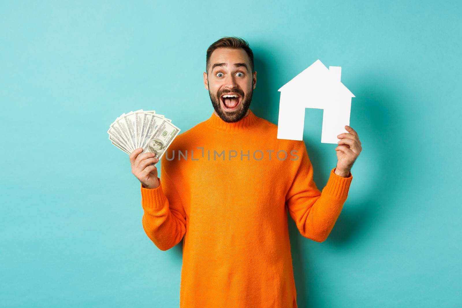 Real estate concept and mortgage concept. Excited man showing dollars and paper maket of house, buying flat, standing over blue background.