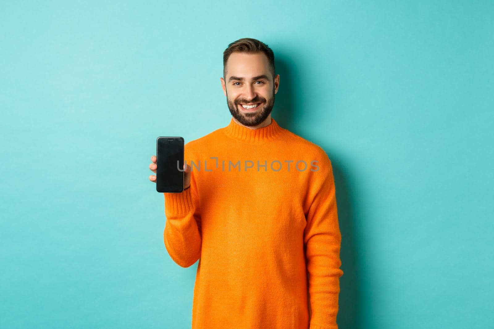 Handsome bearded guy in orange sweater, showing smartphone screen and smiling, showing promo online, turquoise background.