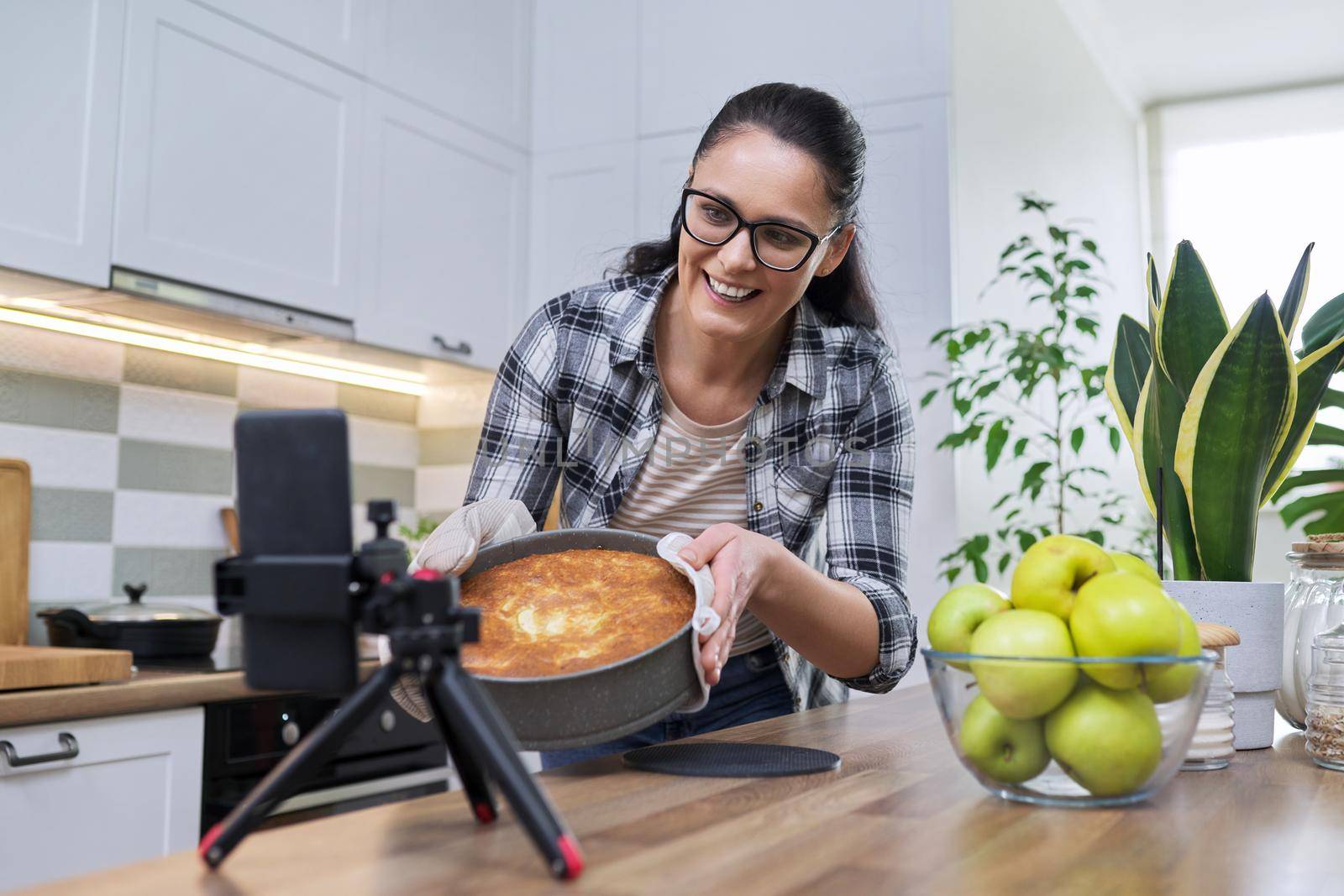 Woman preparing apple pie at home in the kitchen, with a smartphone using a video call by VH-studio