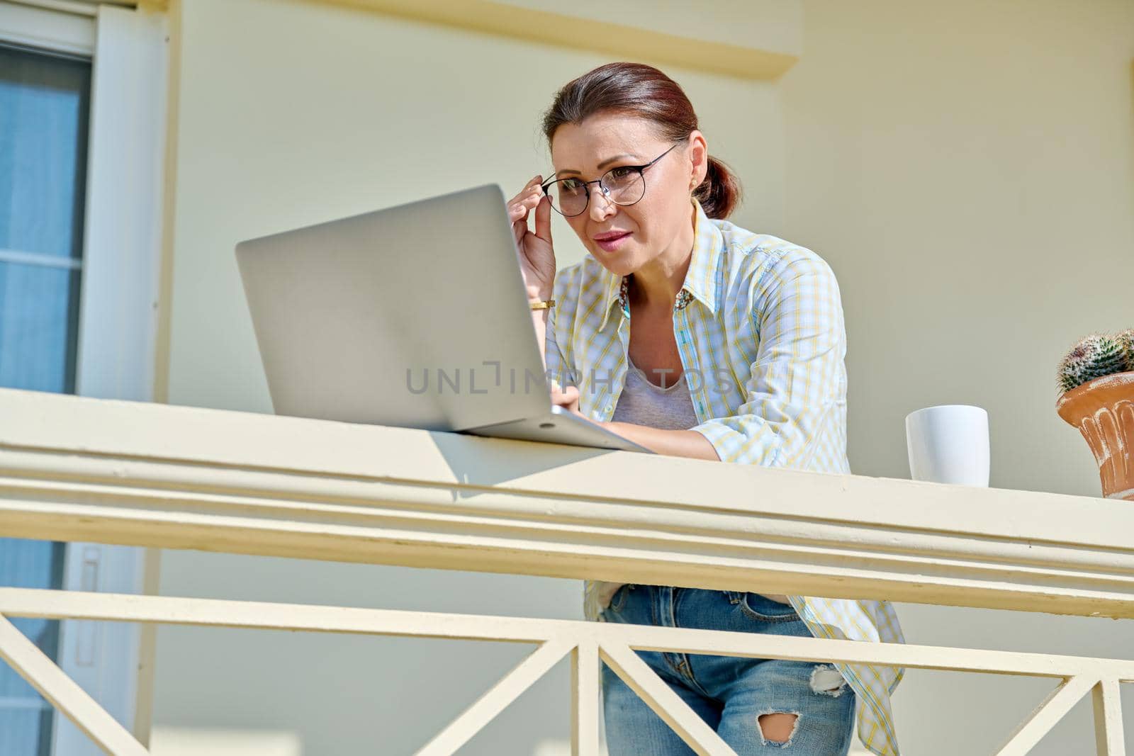 Serious focused middle aged woman with laptop on home balcony. Concentrated 40s mature business woman working remotely. Freelance, technology, work from home office, online training, blog, vlog