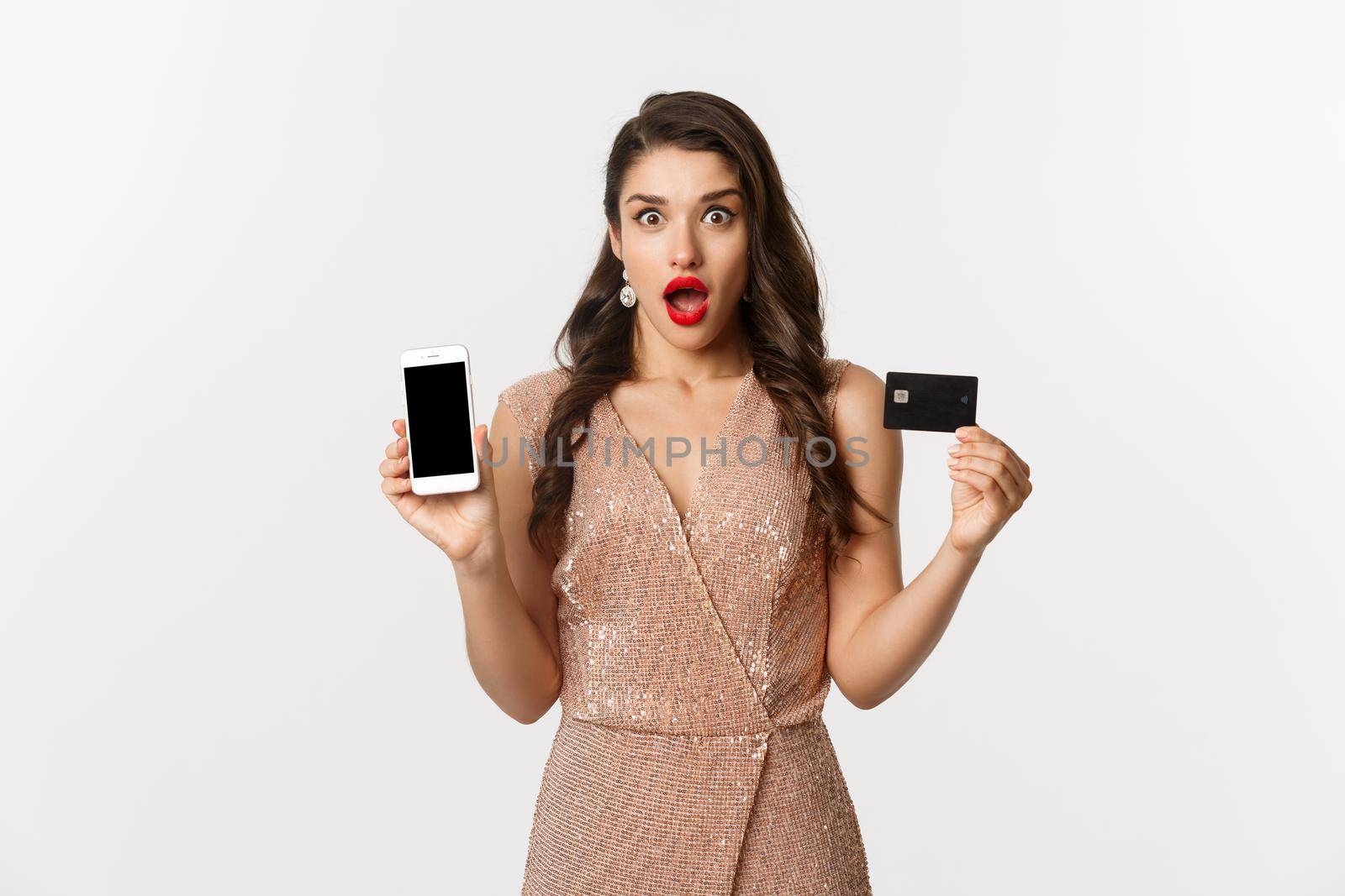 Online shopping and holidays concept. Woman looking amazed and showing credit card with smartphone screen, standing over white background.