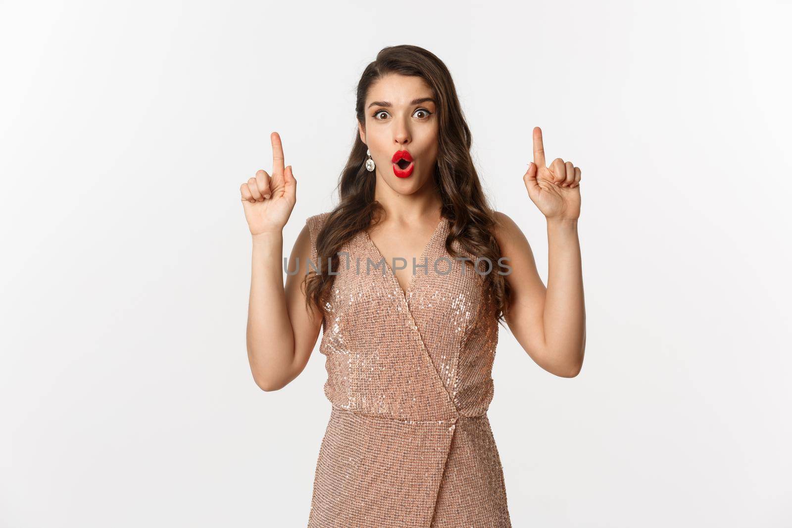 New Year, christmas and celebration concept. Surprised and excited woman in glamour dress, wearing outfit for party, pointing fingers up, showing promo offer, white background.
