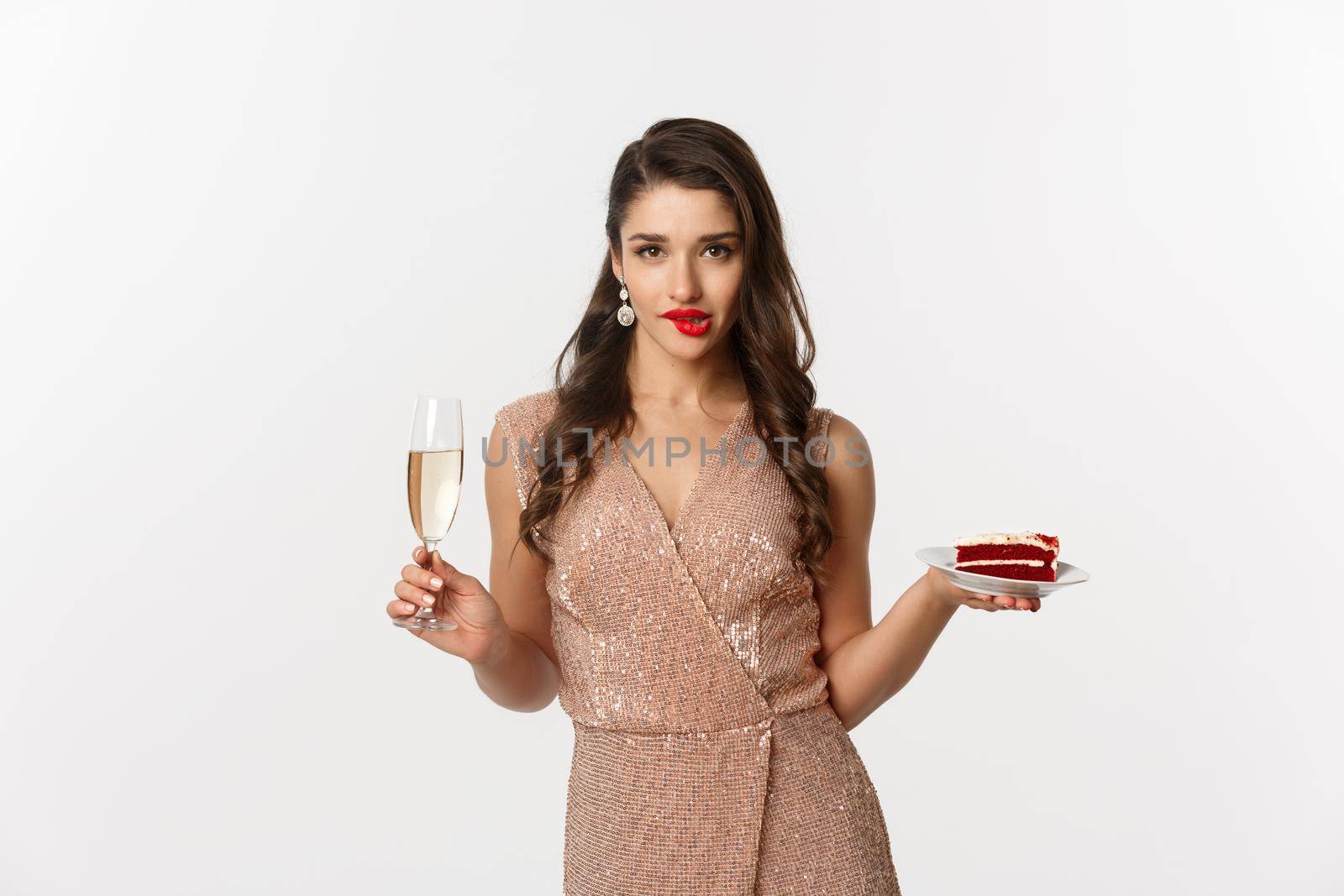 Party and celebration concept. Sexy woman in elegant dress, holding champagne and piece of cake, biting lip and looking seductive at camera.