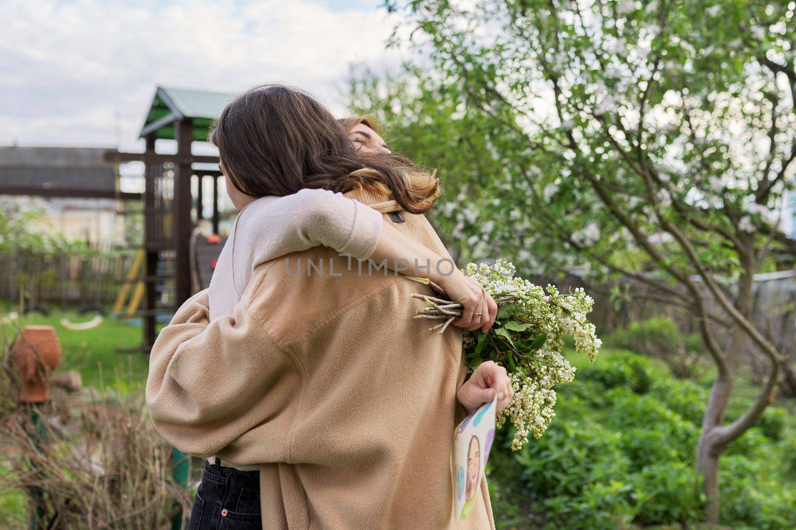 Teenage son congratulating mom on Mother's Day with a bouquet of lilac flowers and a handicraft picture, outdoor in the backyard. Family, holiday, relationship, love, happiness