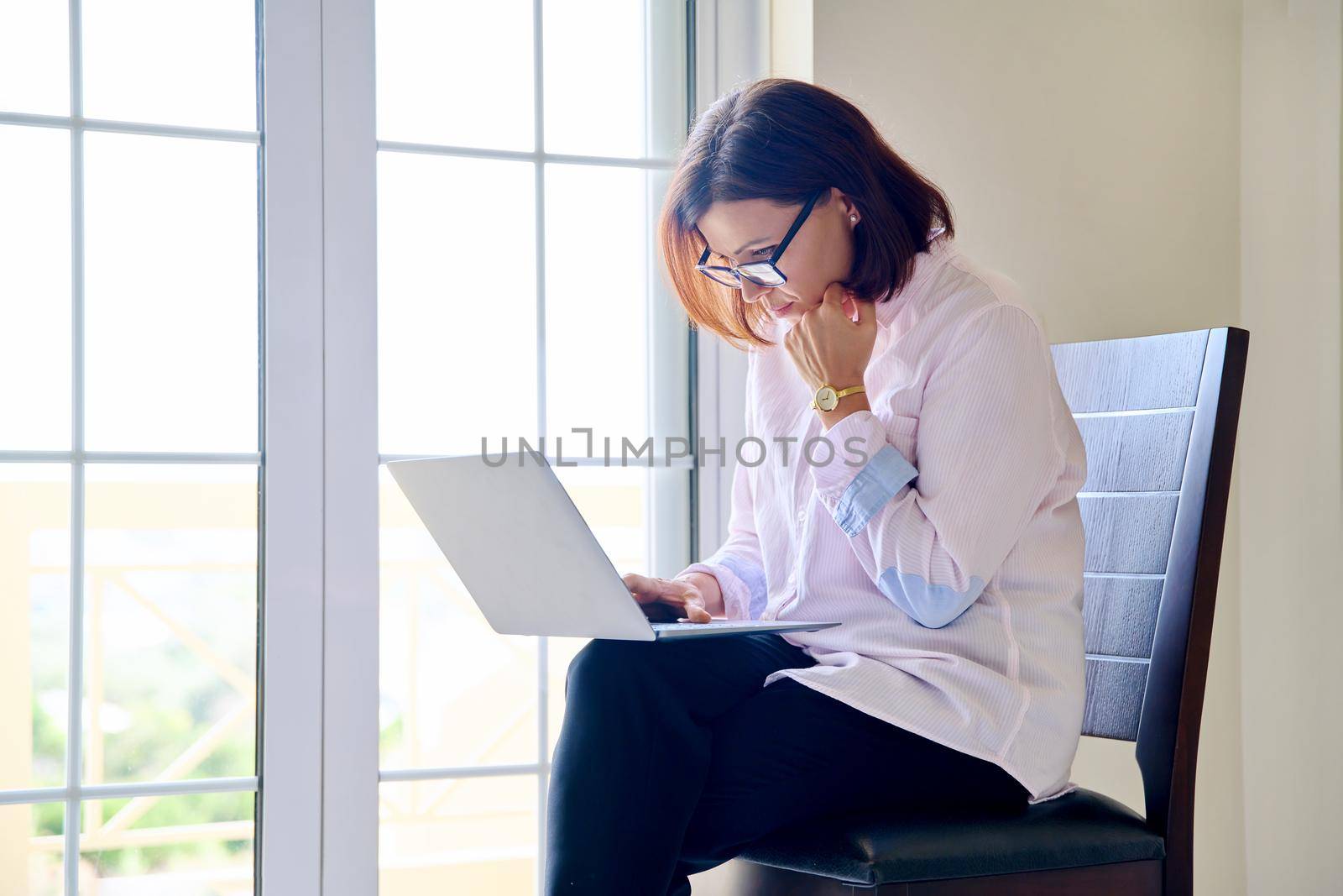 Mature businesswoman in glasses with laptop in her hands near window by VH-studio