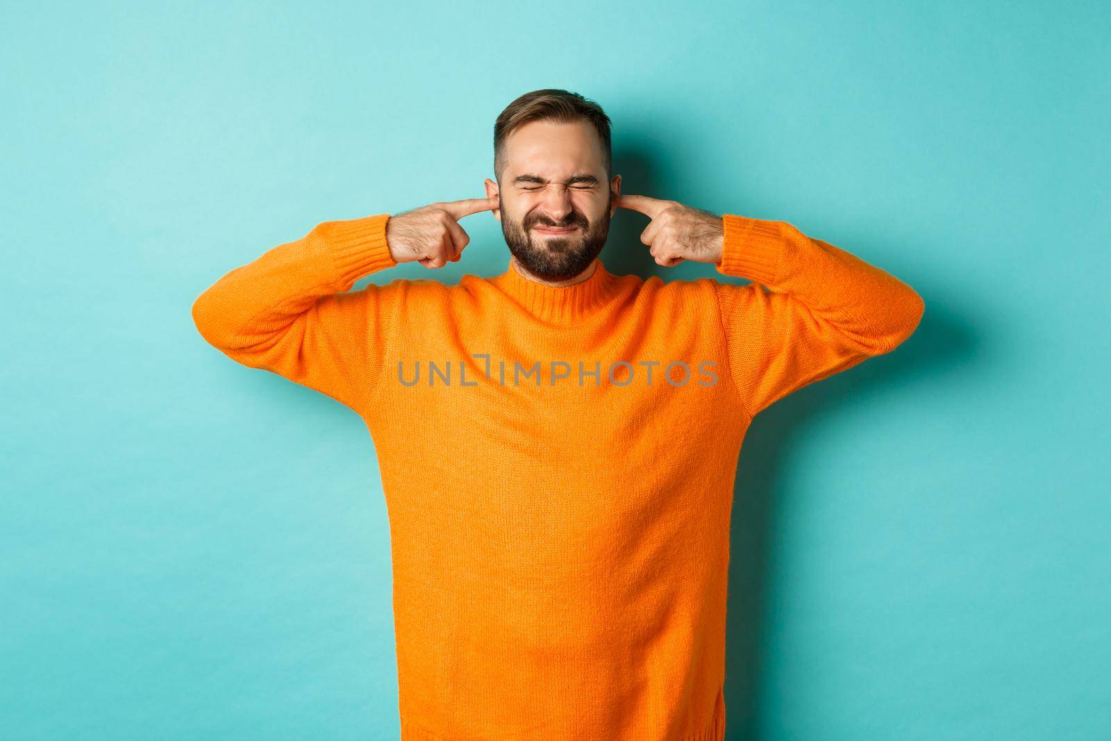 Displeased man shut ears and grimacing, loud neighbours, standing annoyed with noise, light blue background.