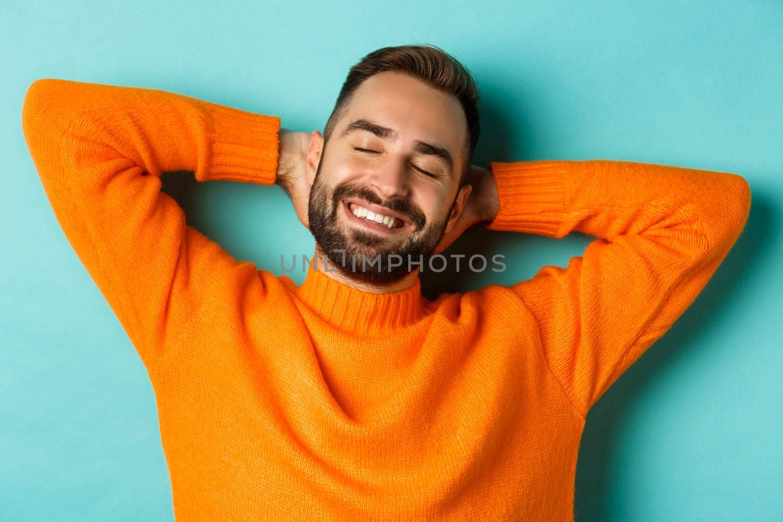 Carefree young man daydreaming, resting with hands behind head and closed eyes, relaxing over turquoise background in orange sweater.