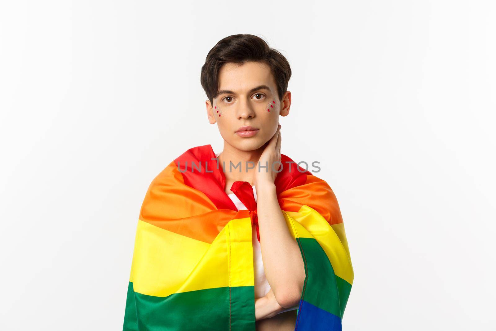 Waist-up shot of beautiful gay man with glitter on face, wrap himself with rainbow lgbt flag and touching face gently, looking at camera, white background.