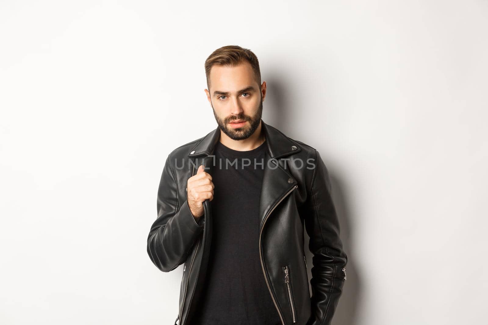 Handsome bearded man expressing confidence, touching his leather jacket and looking self-assured, white background.