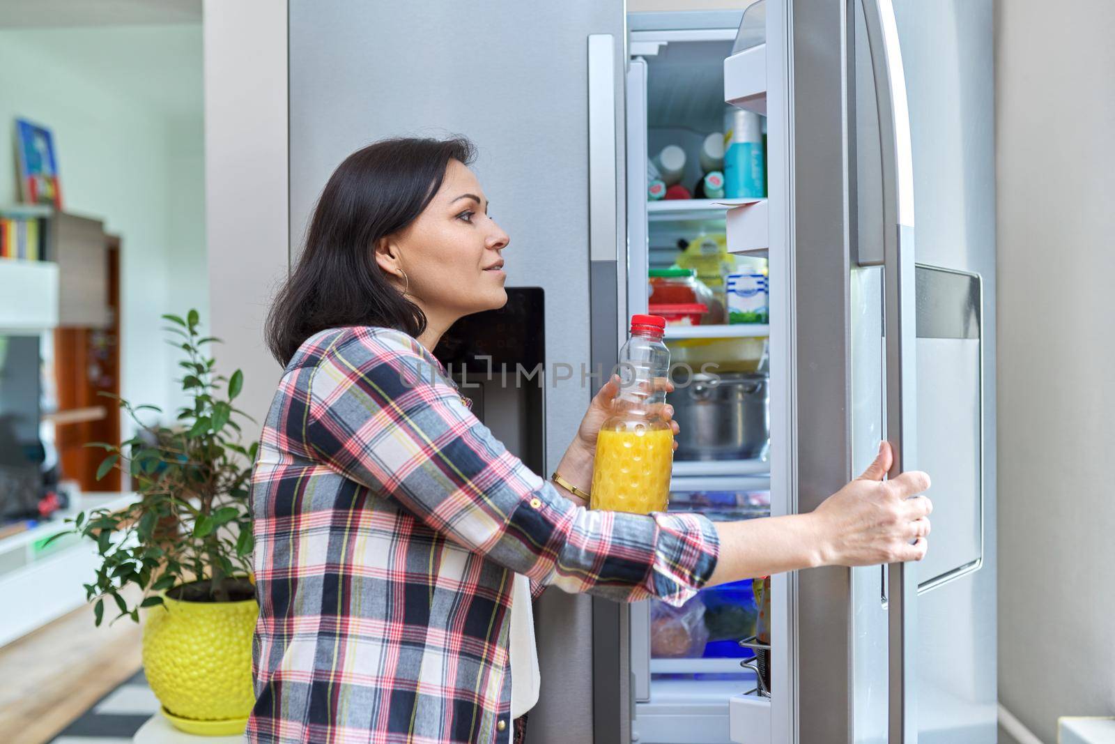 A woman opens the refrigerator at home in the kitchen, holding a bottle of orange juice. by VH-studio