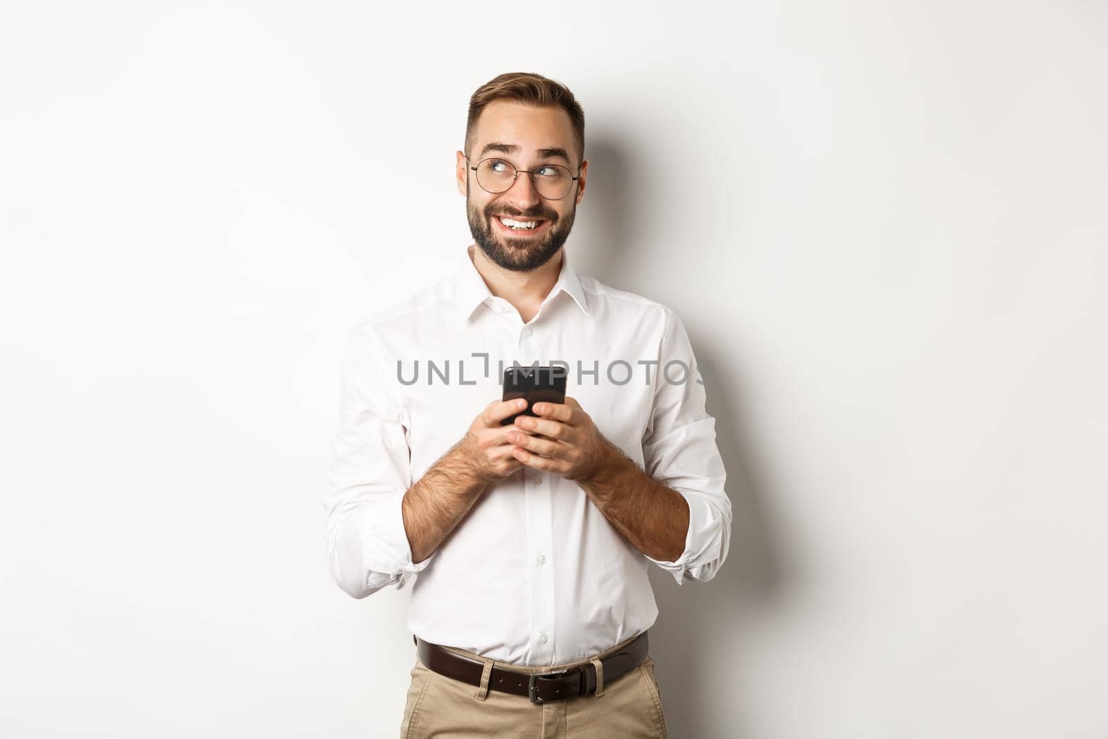 Thouthful handsome manager using mobile phone and thinking about answering message, looking at upper left corner and smiling, standing over white background.