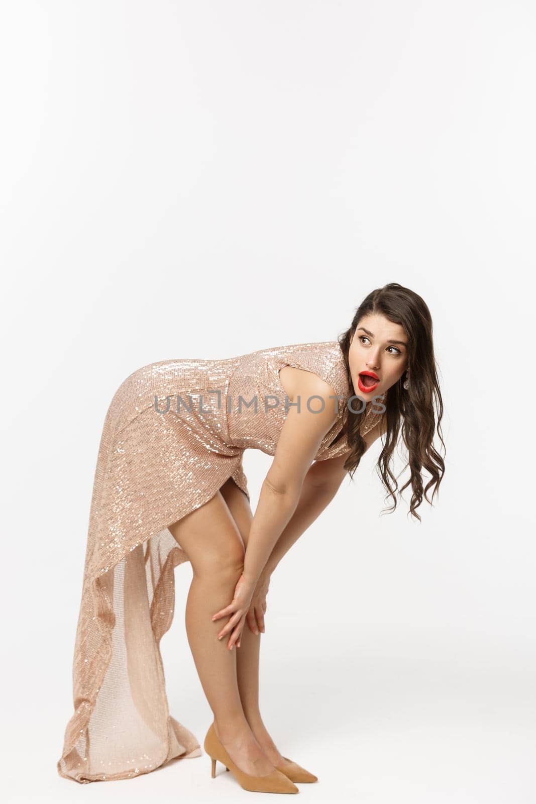 Christmas party and celebration concept. Full length of sensual brunette in luxury dress, bend down and looking left with surprised face, white background.