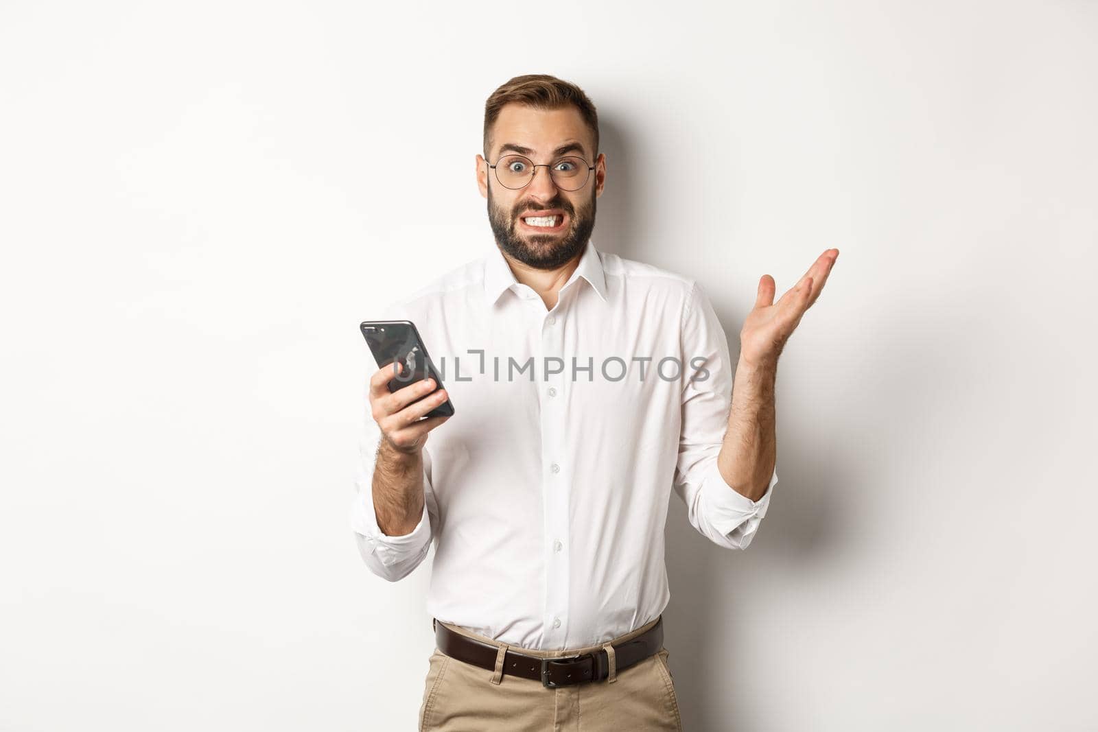 Confused businessman reading strange message on mobile phone, looking annoyed, standing over white background.