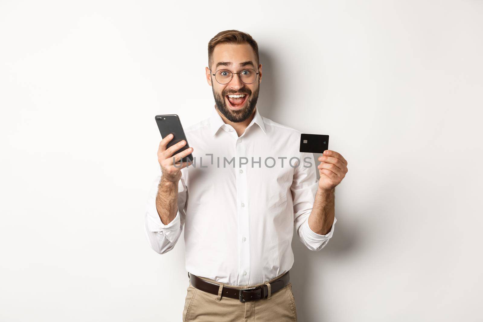 Business and online payment. Excited man paying with mobile phone and credit card, smiling amazed, standing over white background by Benzoix