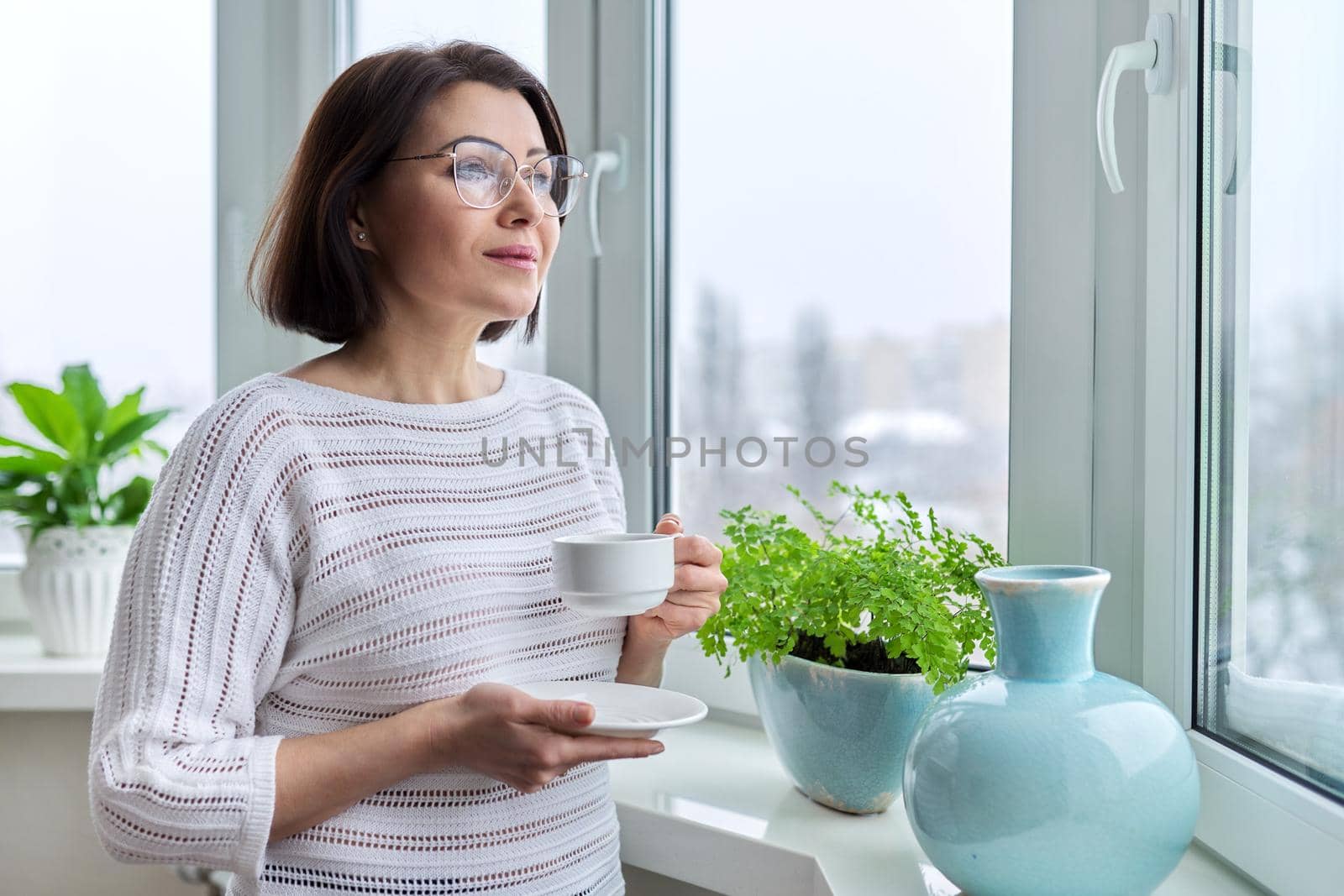 Middle aged beautiful woman with glasses with cup of coffee at home in winter autumn season by VH-studio