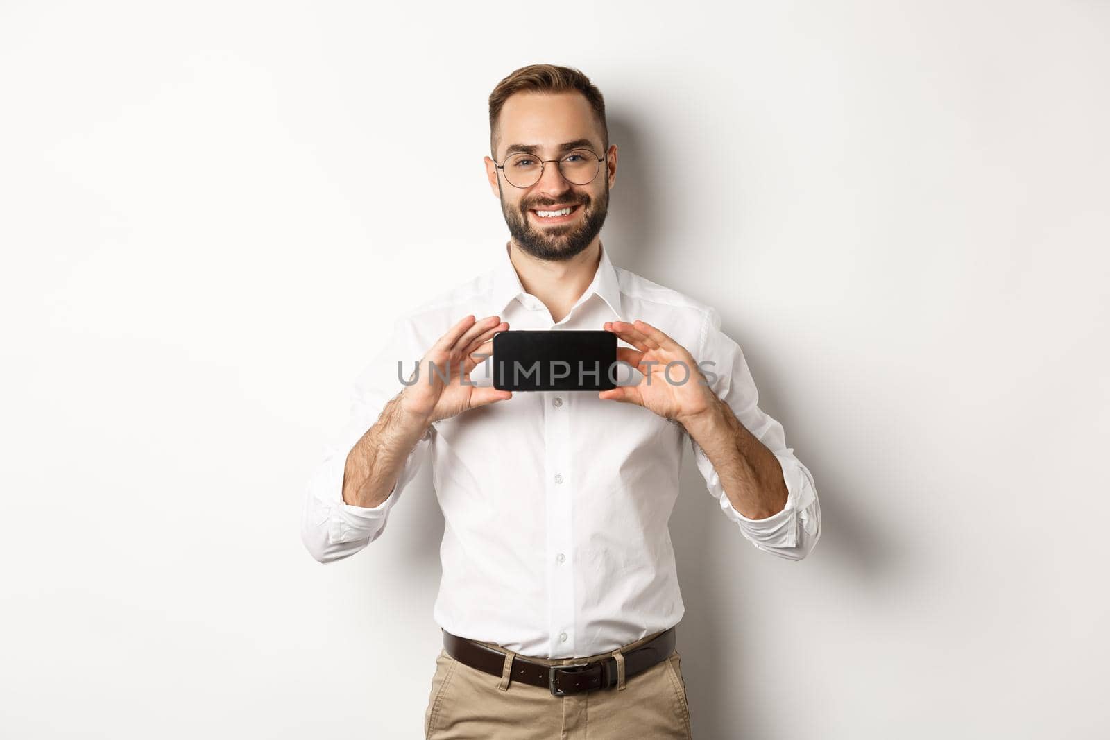 Handsome bearded man in glasses, demonstrating mobile phone application, showing smartphone screen, standing over white background.