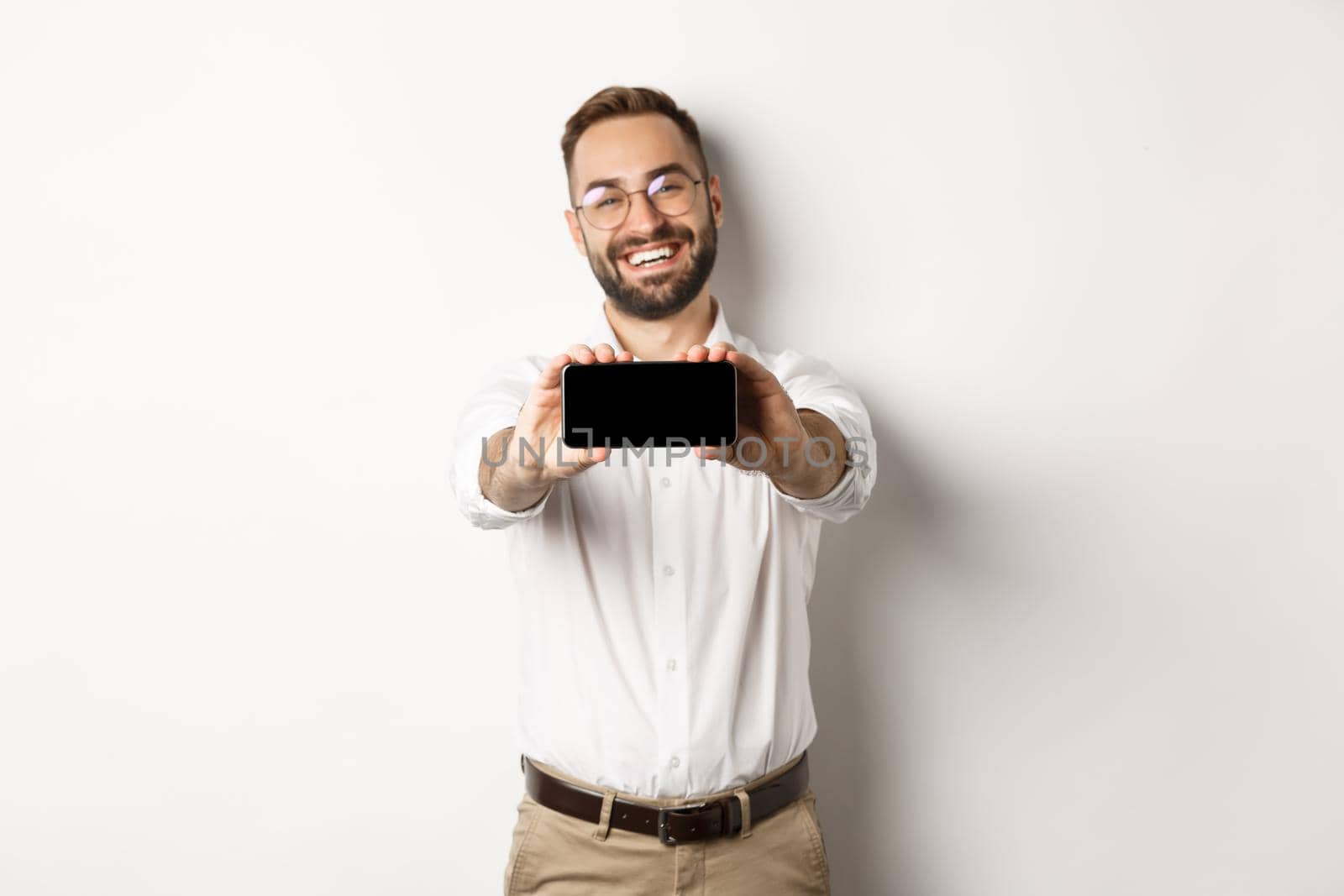 Happy business man showing mobile screen, holding phone horizontally, standing satisfied against white background by Benzoix