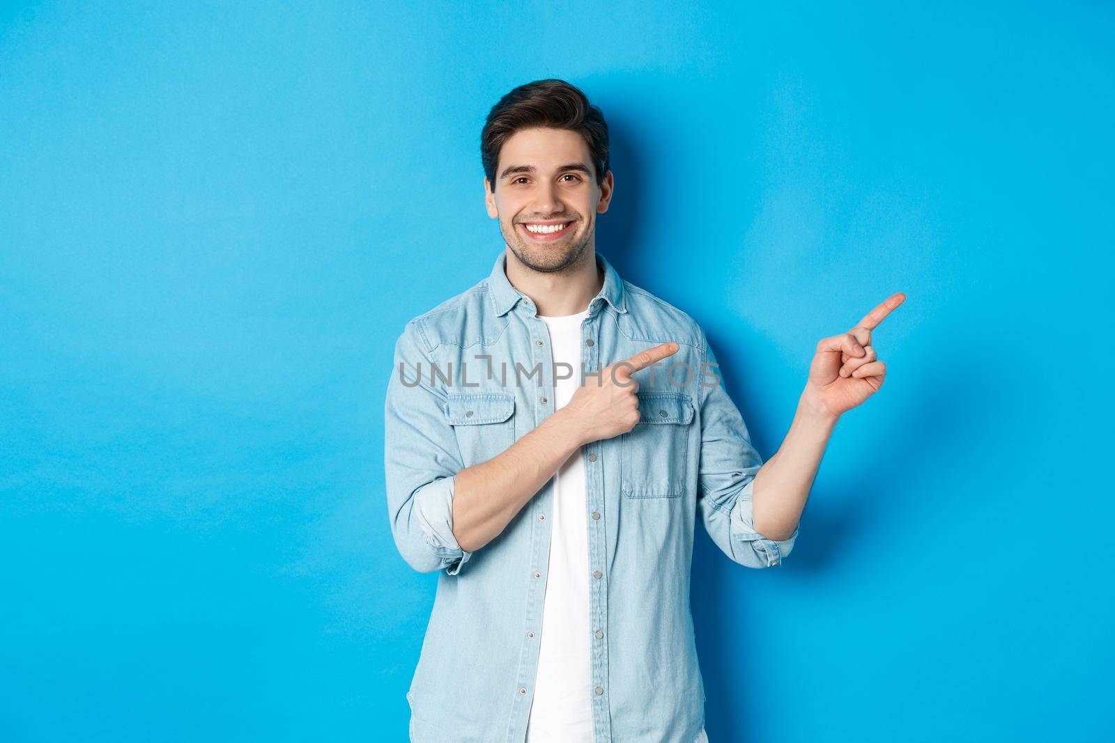 Portrait of attractive adult man smiling, pointing fingers right at logo or banner, showing advertisement against blue background.