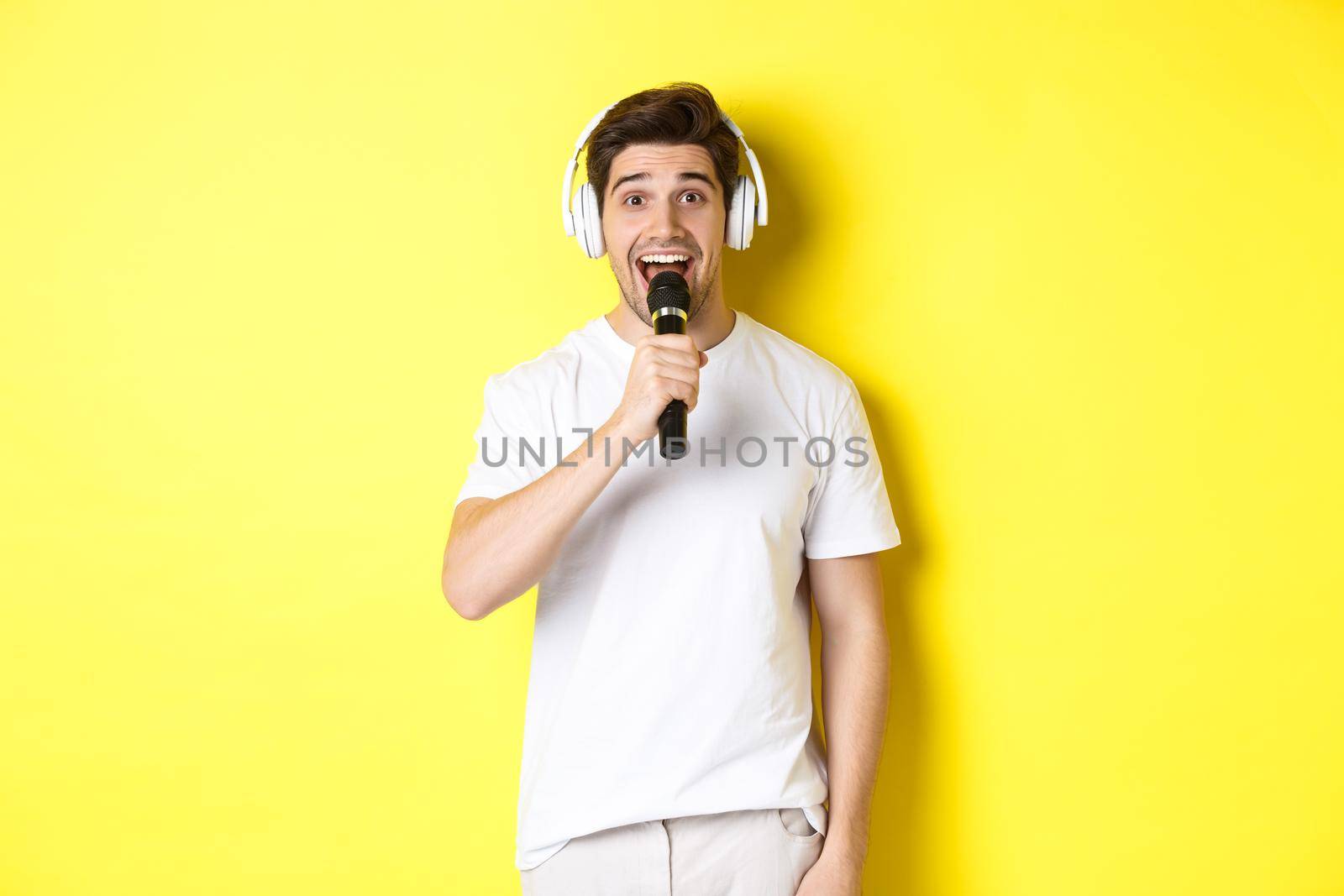 Man in headphones holding microphone, singing karaoke song, standing over yellow background in white clothes by Benzoix