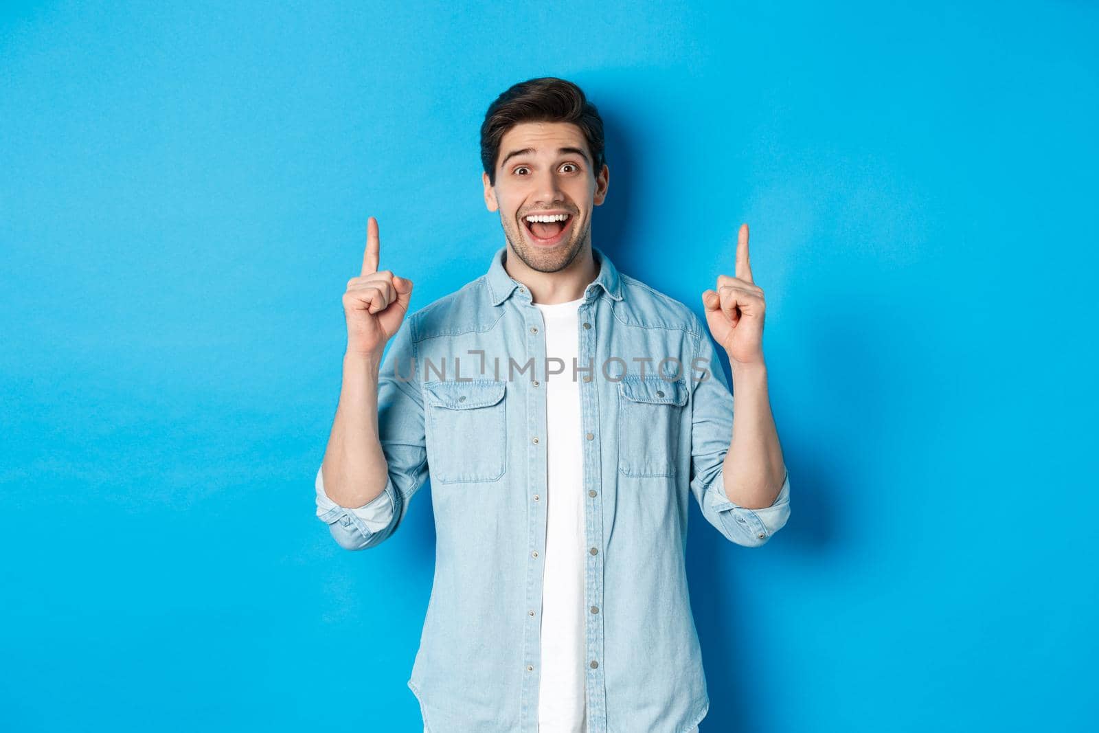 Portrait of happy 25s guy with beard, pointing fingers up and smiling, showing advertisement, standing against blue background.