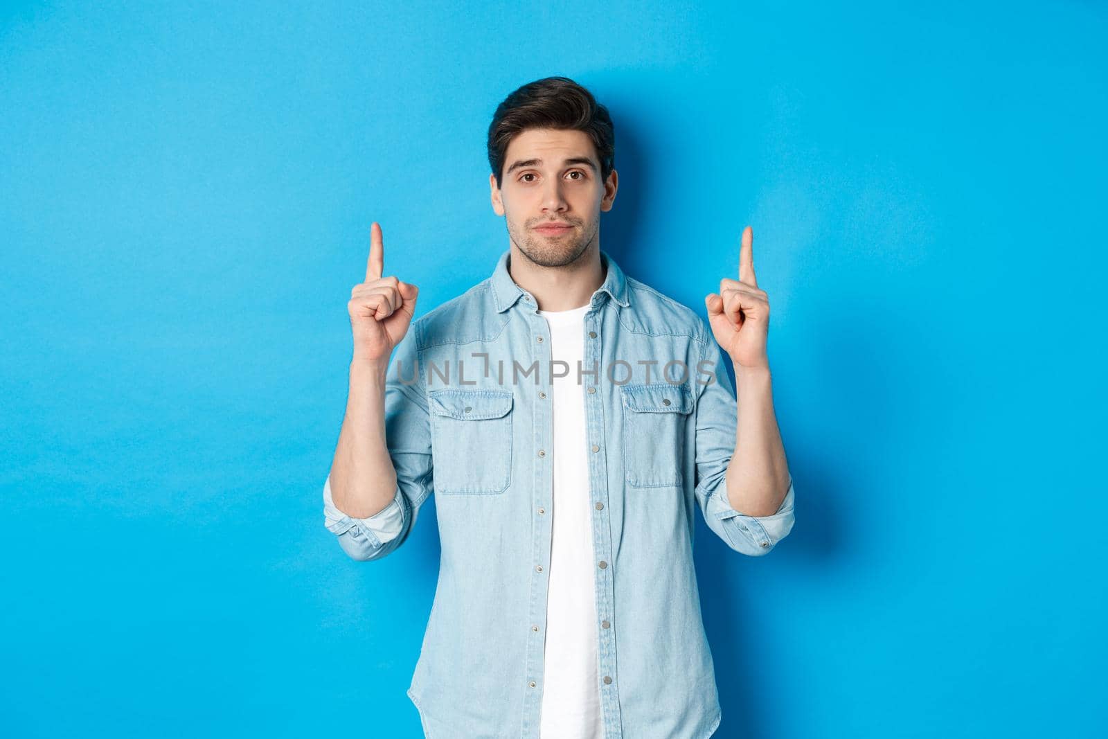 Image of calm handsome man showing you promo offer, pointing fingers up at copy space, standing against blue background.