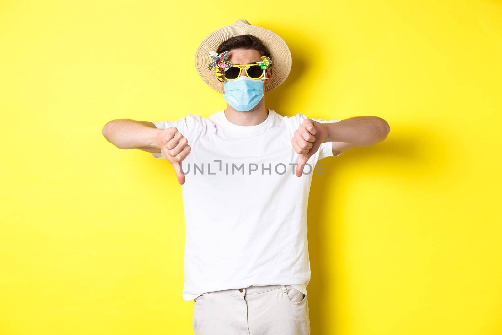 Concept of covid, vacation and tourism. Disappointed tourist complaining on lockdown during pandemic, wearing medical mask and sunglasses, showing thumbs down.