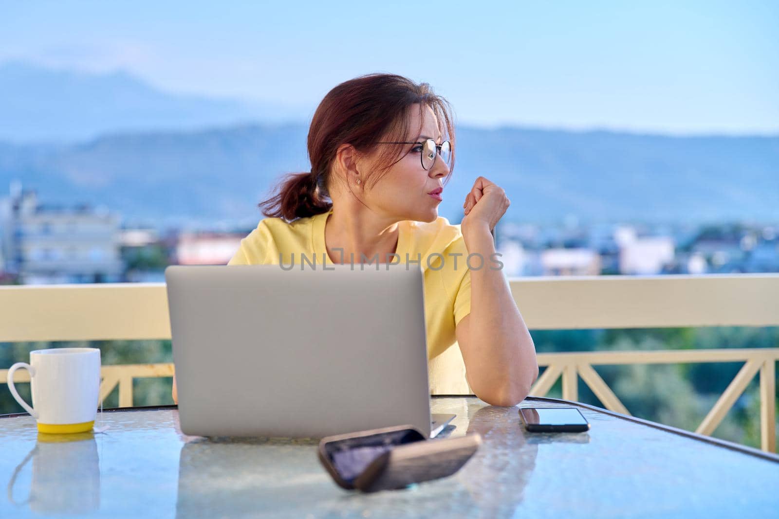 Serious middle aged woman with laptop at table on home outdoor balcony by VH-studio