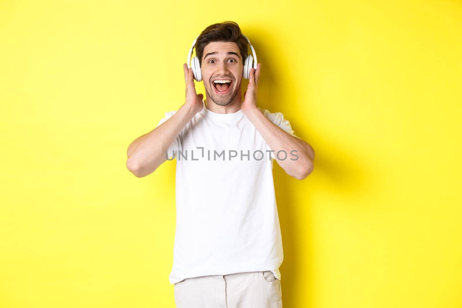 Man in headphones looking surprised and happy, listening awesome song, standing over yellow background by Benzoix