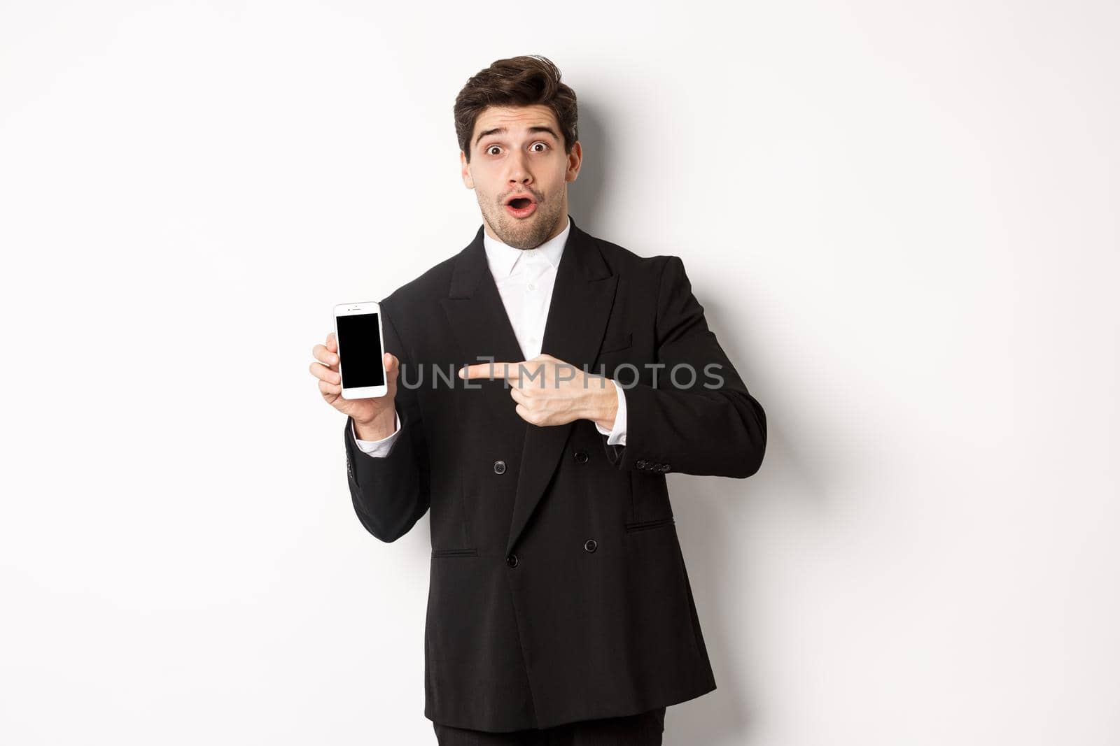 Portrait of attractive businessman in black suit, looking surprised and pointing finger at smartphone sreen, standing over white background by Benzoix