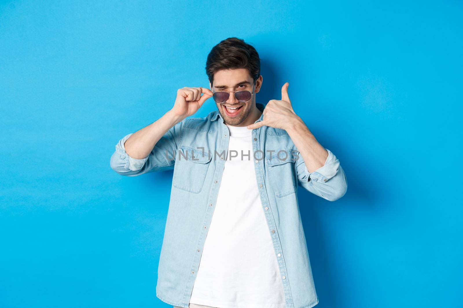 Handsome macho man in sunglasses flirting with you, make phone sign and winking, call me gesture, standing over blue background by Benzoix